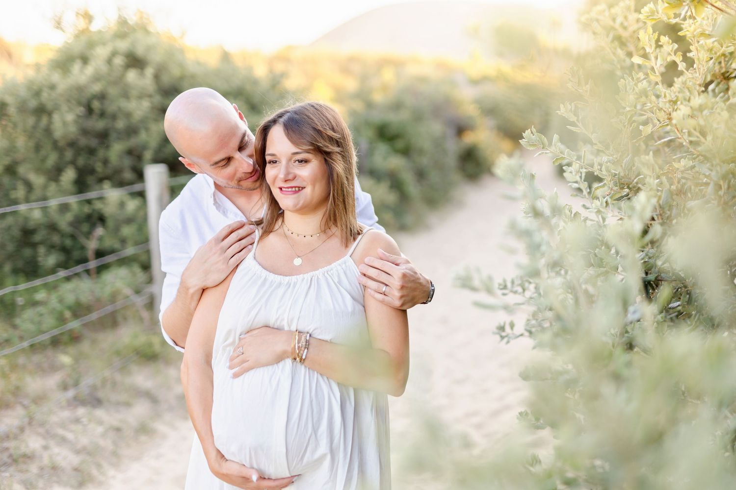 A couple embraces on a sunlit path in nature, wearing light colored clothing for a maternity photoshoot.