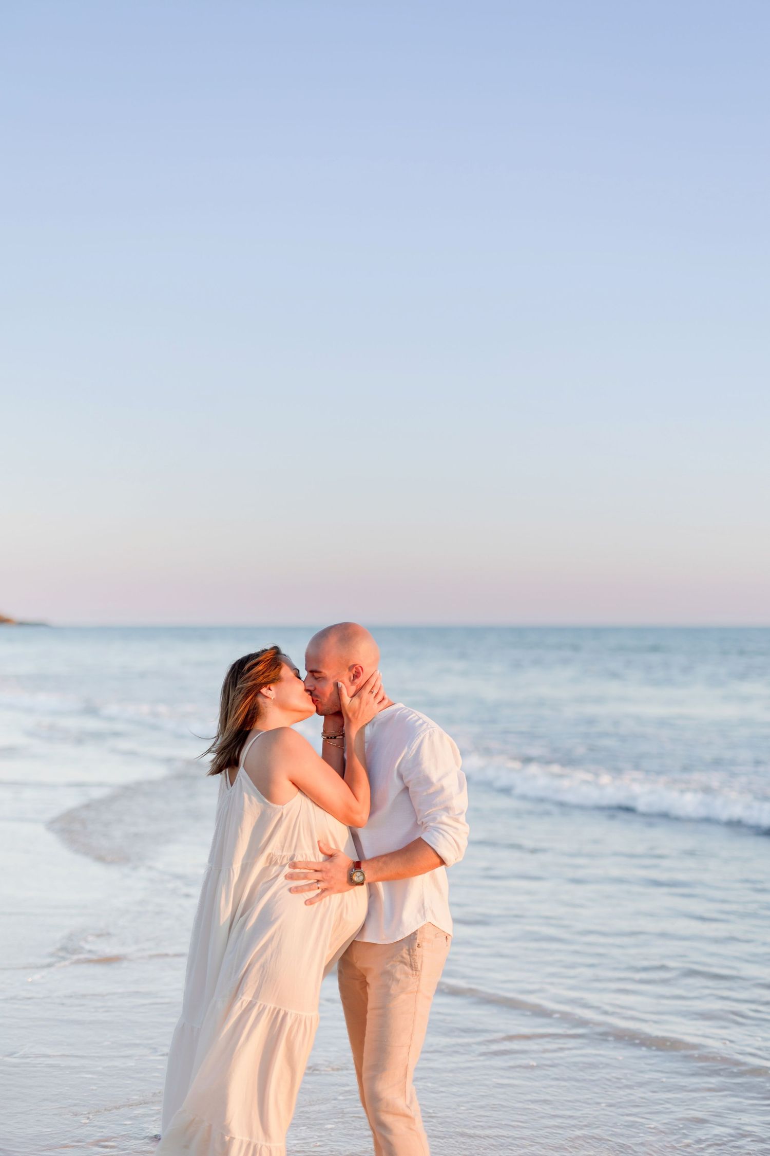Couple in white beach attire sharing a romantic moment by the ocean at sunset.