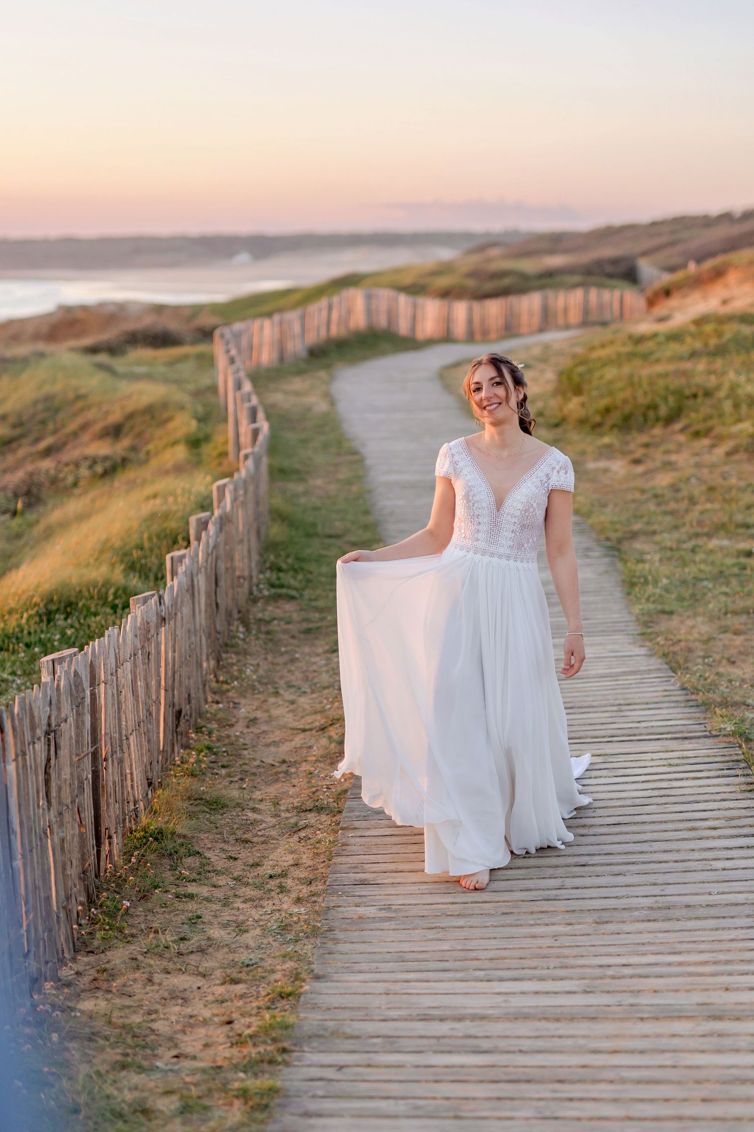 Bride in flowing white dress poses on wooden boardwalk by ocean at sunset.