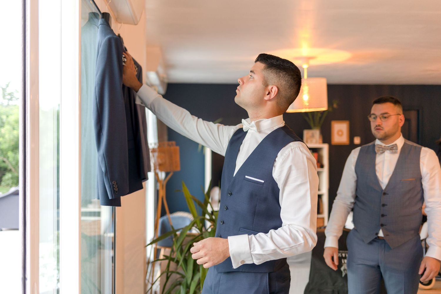 Wedding preparations with groomsmen adjusting suits hanging by window with decorative plants nearby.