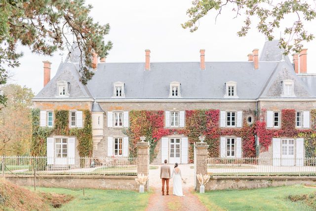 Romantic newlyweds stroll hand in hand beside a grand stone mansion with ivy-covered walls.