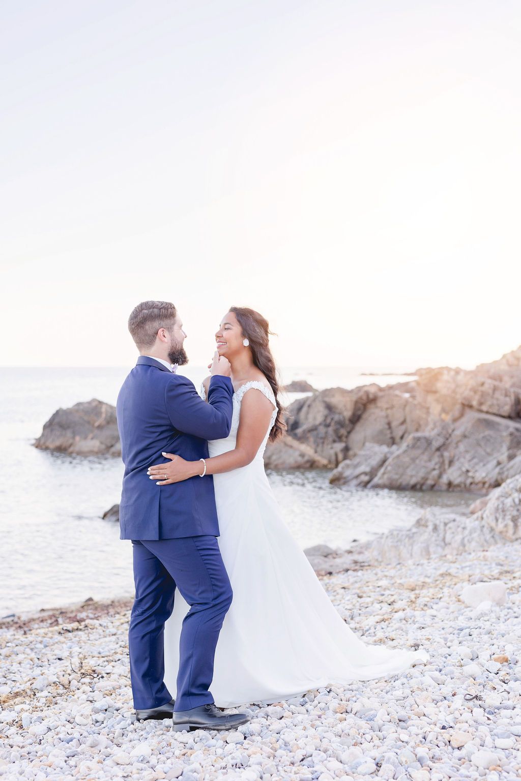 A tender wedding moment photographed on a pebble beach with ocean rocks in background.