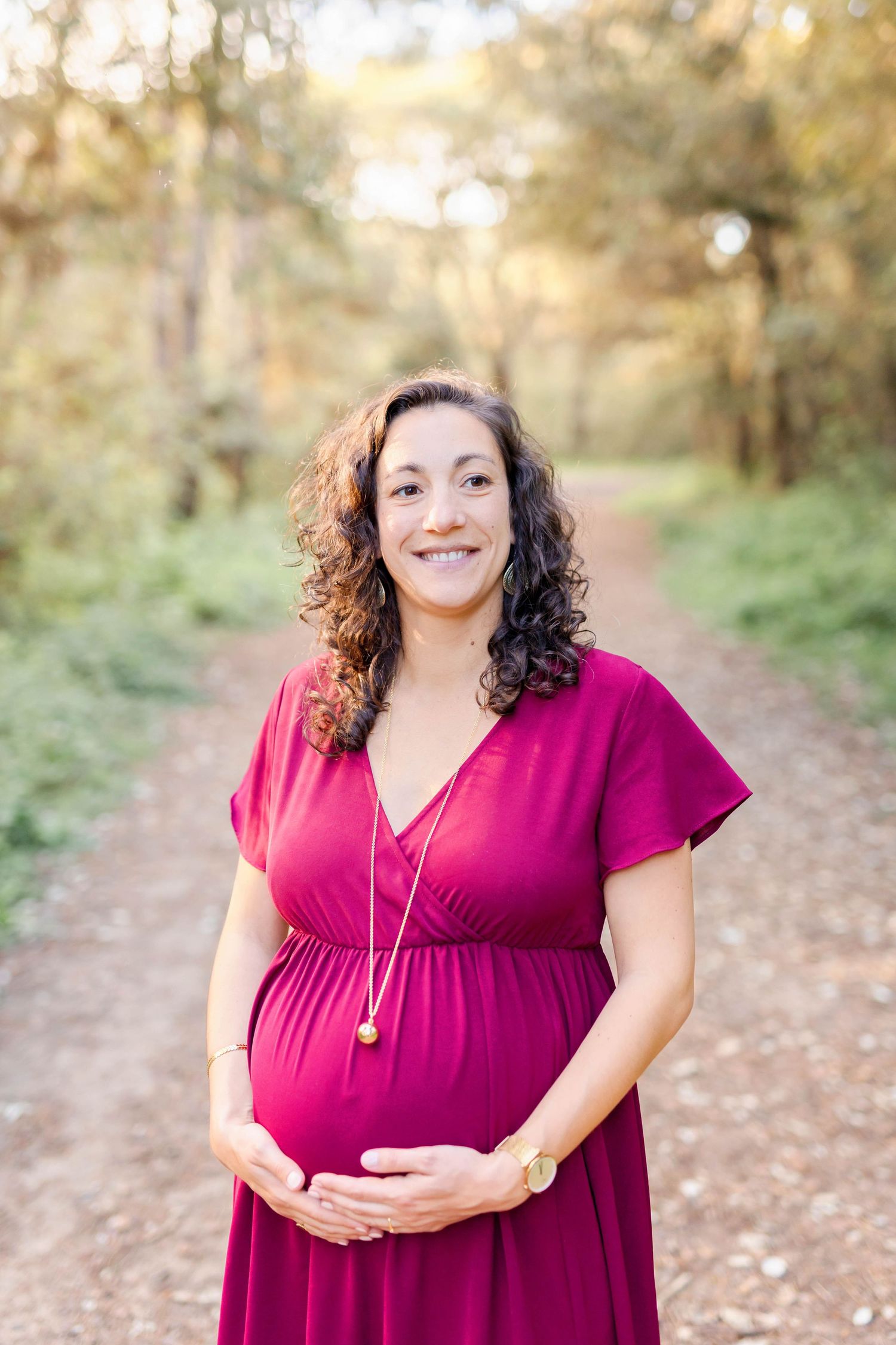 Woman in burgundy dress poses for maternity photos outdoors with natural lighting and greenery.