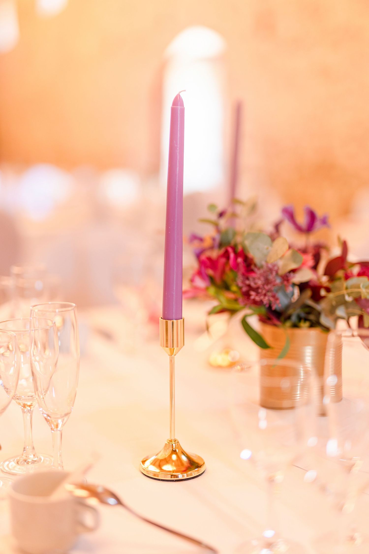 Purple taper candle in gold holder with pink floral arrangement on elegant table setting.