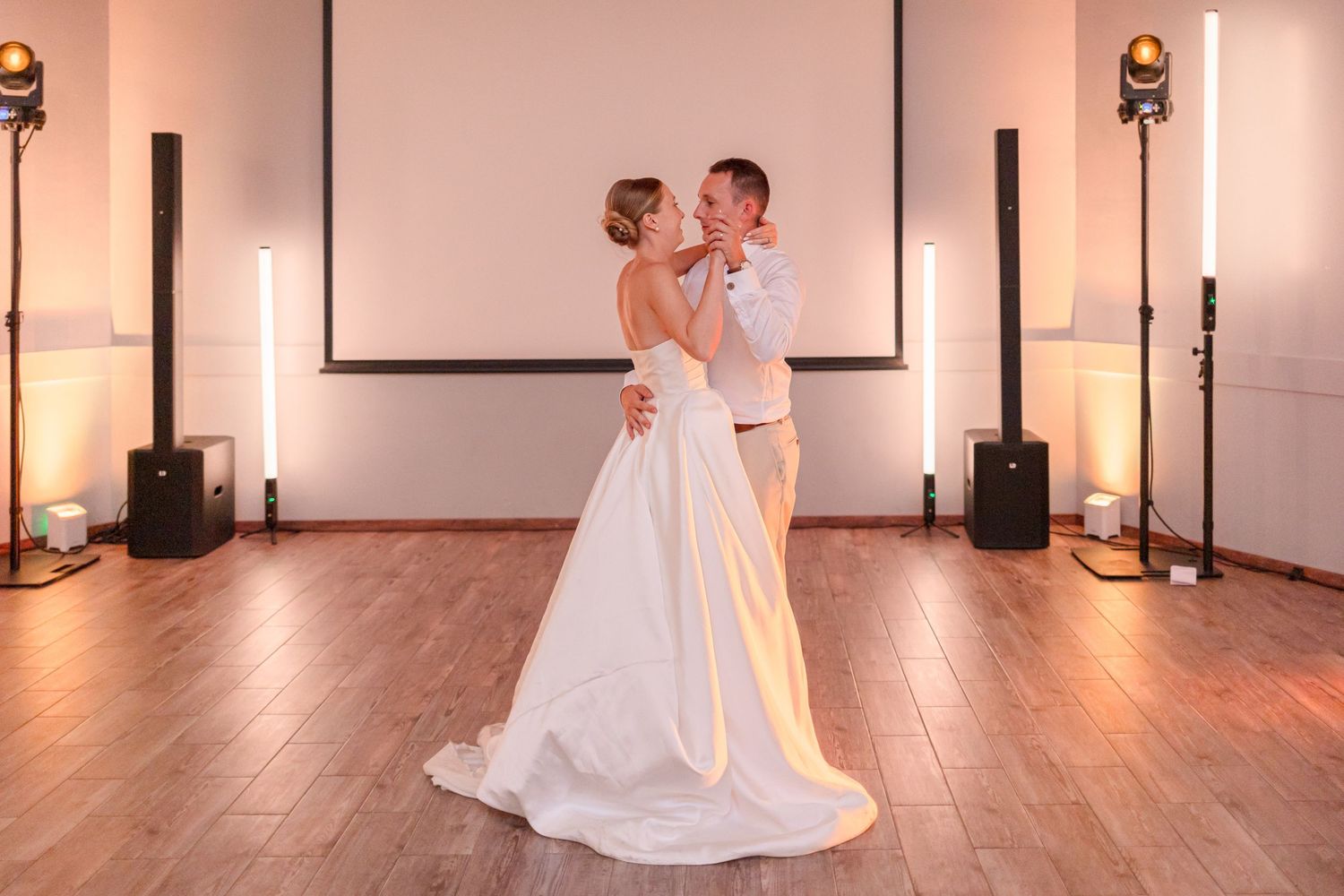 Couple shares an intimate dance moment on wooden floor with projection screen and lighting in background.