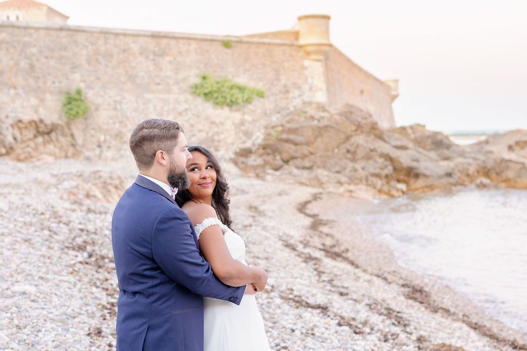 A couple shares a tender moment on a beach with an old fortress wall in the background.
