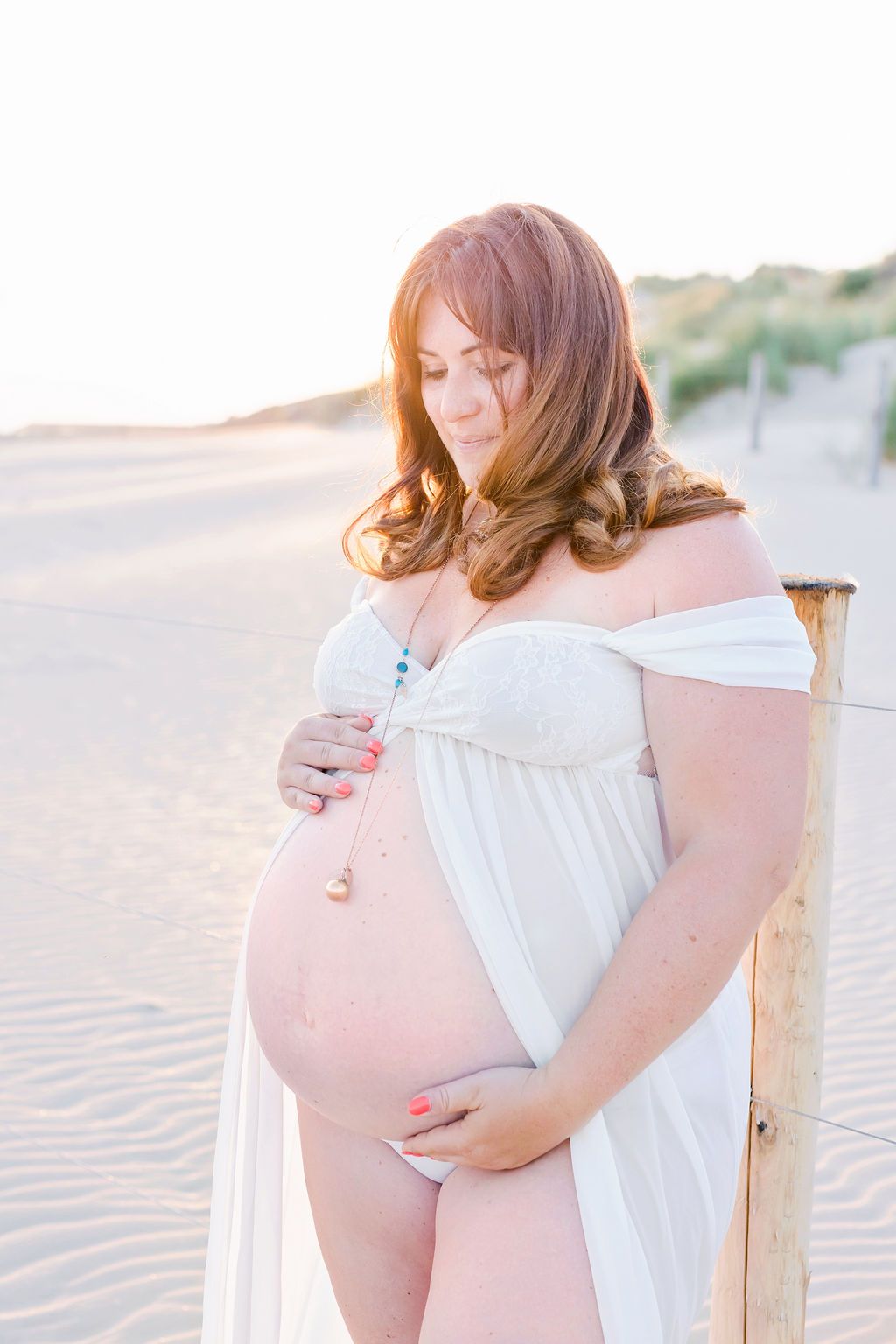 Ethereal maternity portrait on beach with off-shoulder white gown in soft lighting.