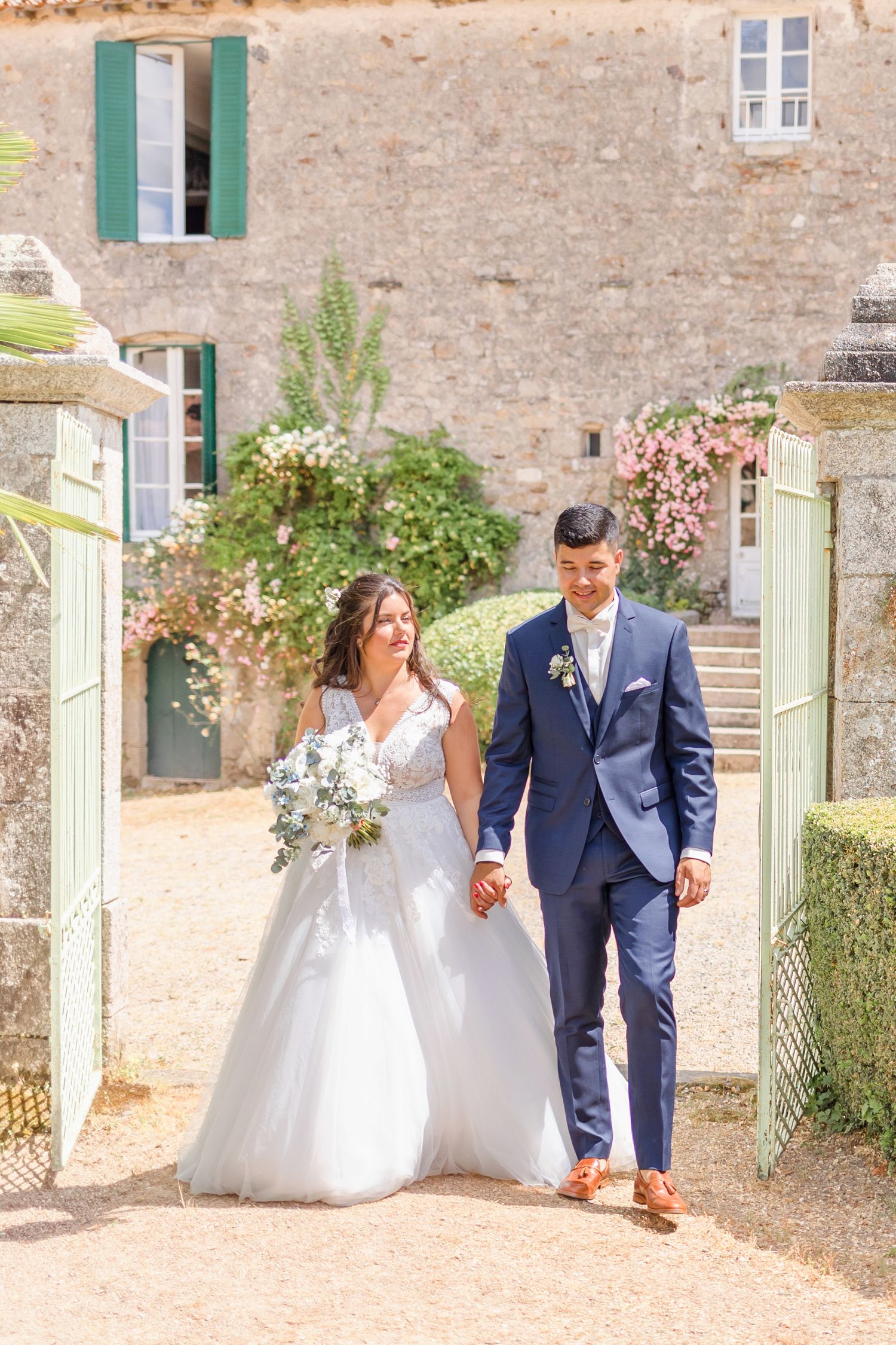 Newlyweds stroll along garden path beside Mediterranean villa with green shutters.