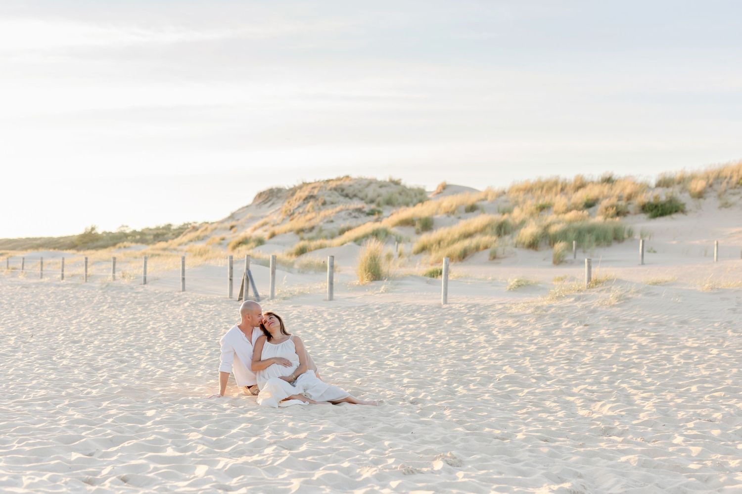 Two people sit together on a beach with sand dunes and fence posts in the background.