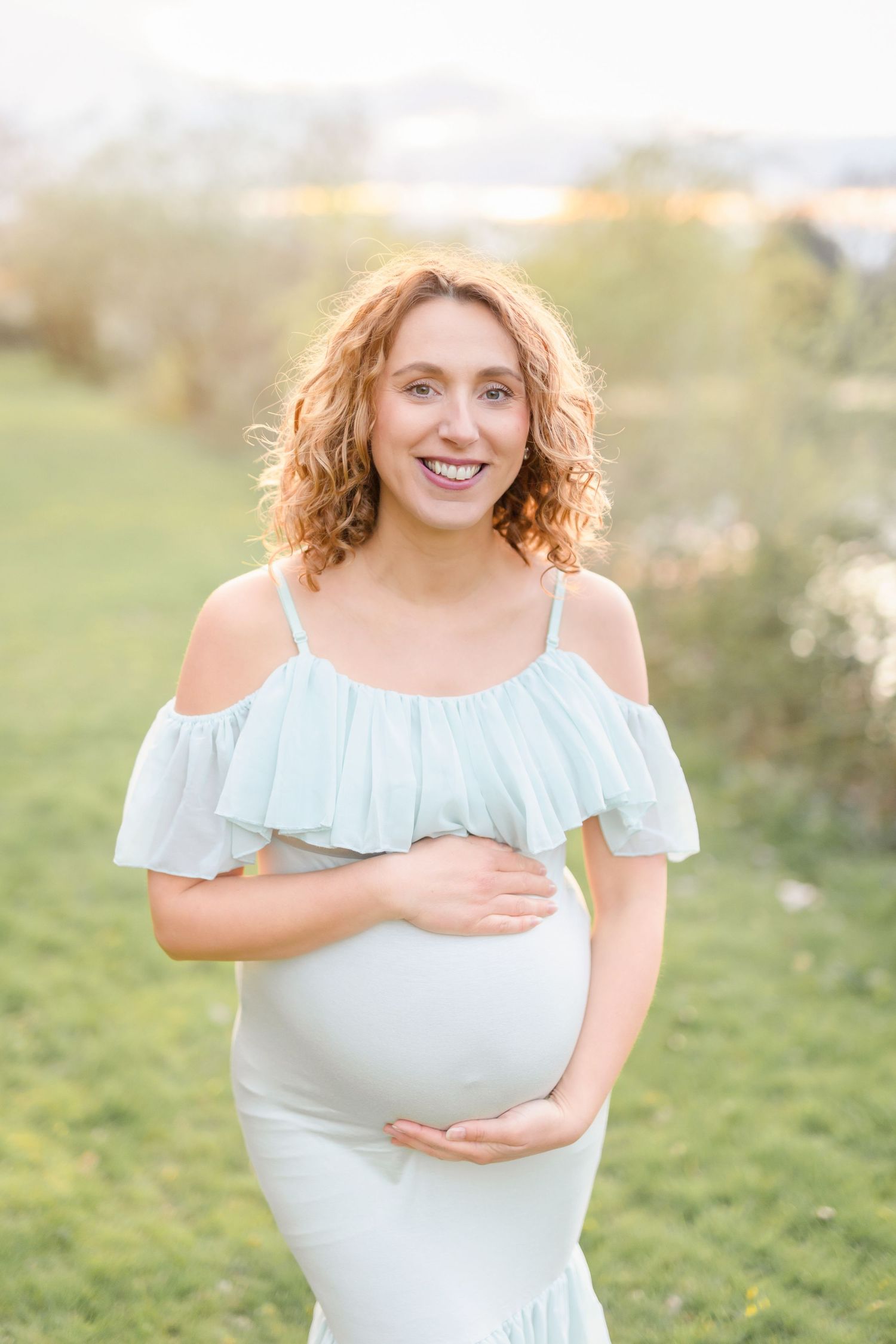 Maternity photo featuring a flowing pale blue cold-shoulder dress in a sunlit outdoor setting.