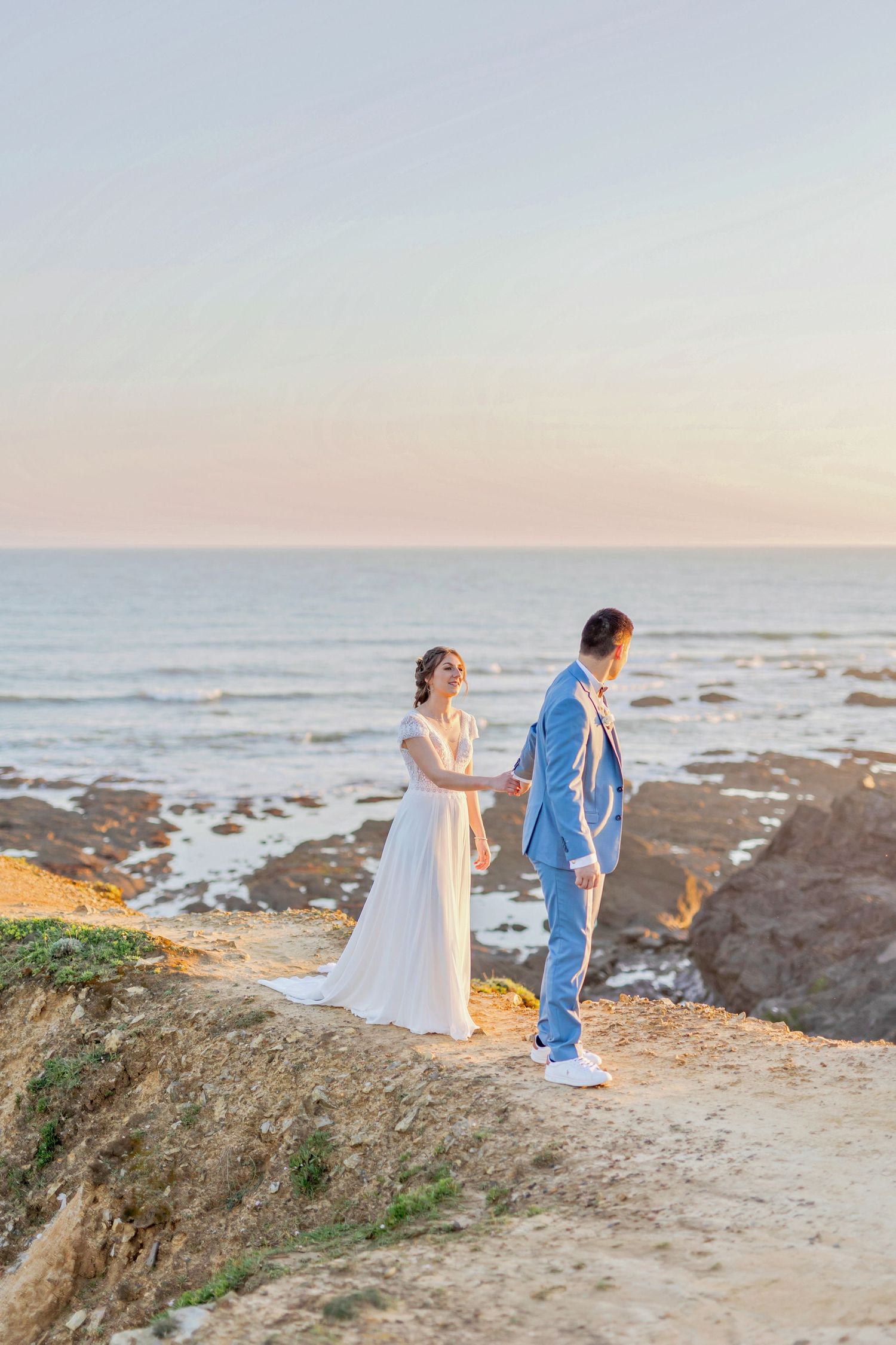 Bride in flowing white dress and groom in blue suit walking along rocky coastline.