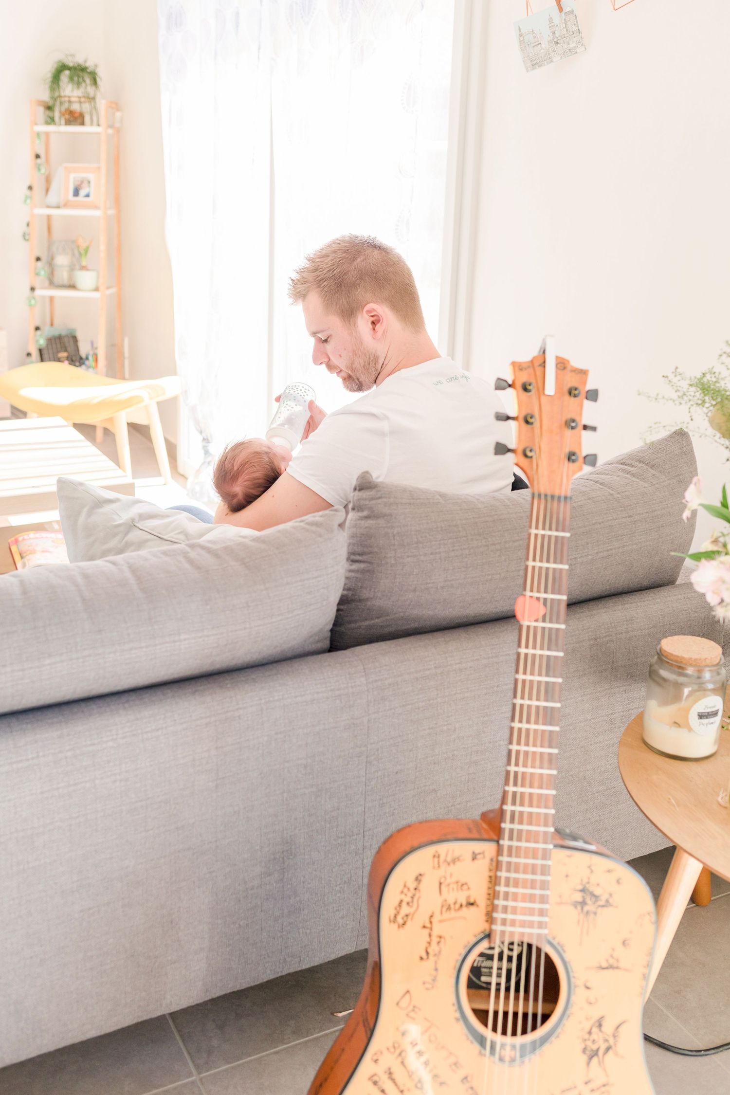 An acoustic guitar rests on a gray couch in a bright, sunlit living room.