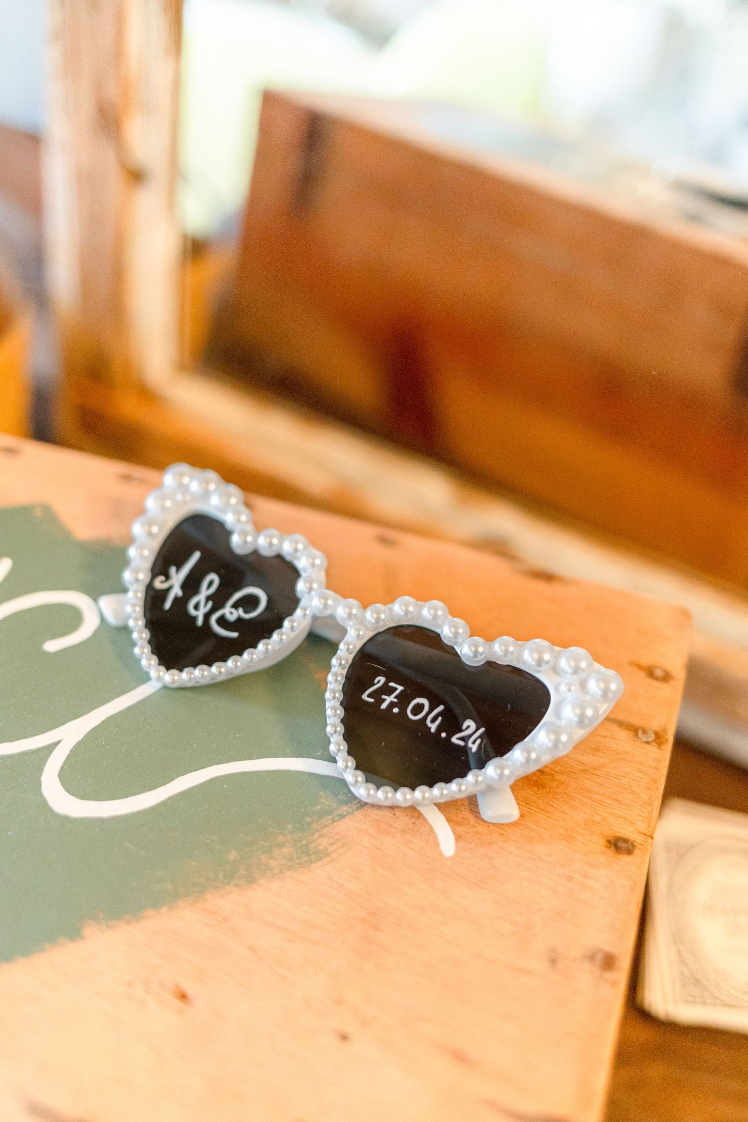 Heart-shaped sunglasses decorated with pearls displaying text on black lenses sit on a wooden surface.