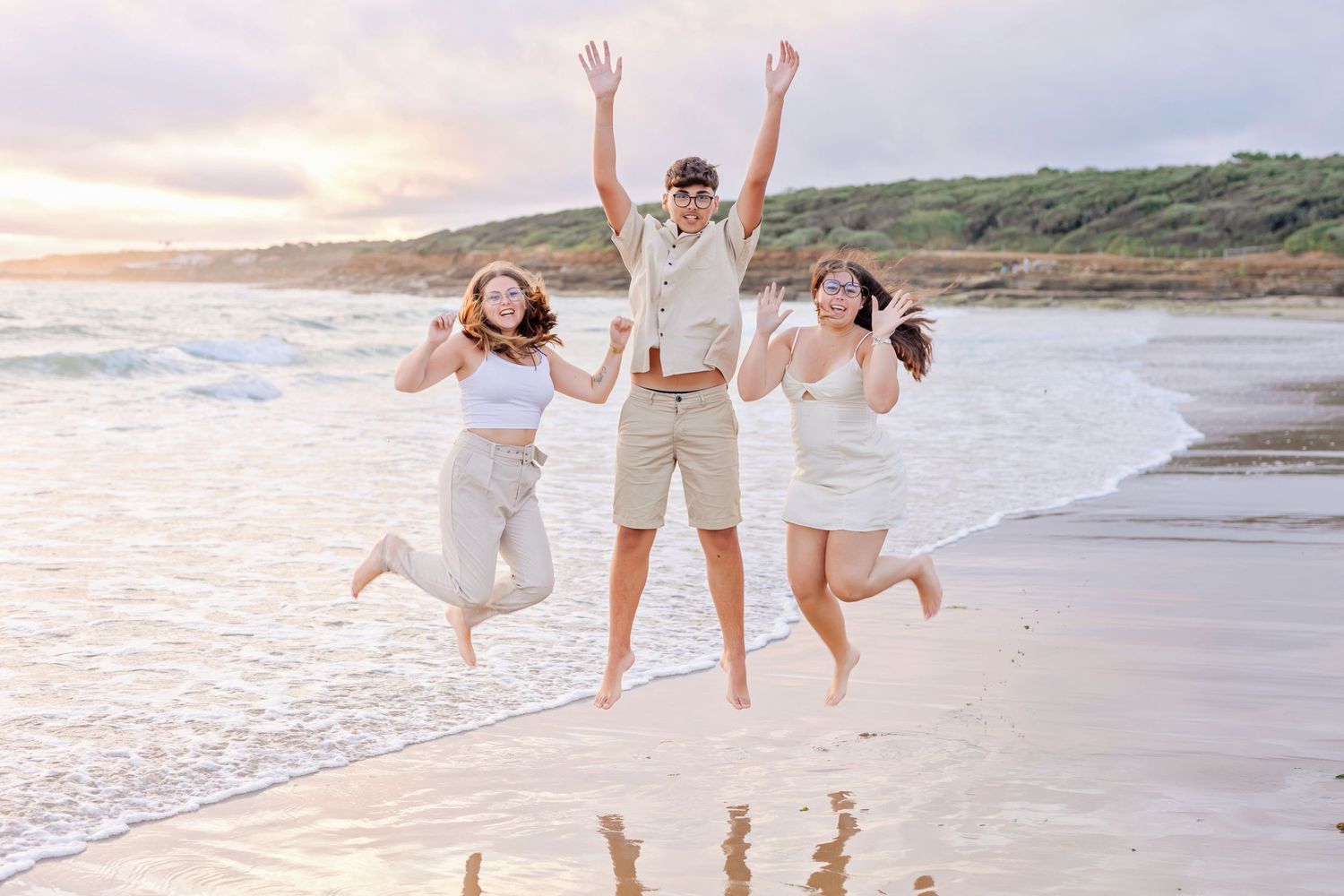 Three people jumping joyfully on a beach at sunset wearing white and beige outfits.