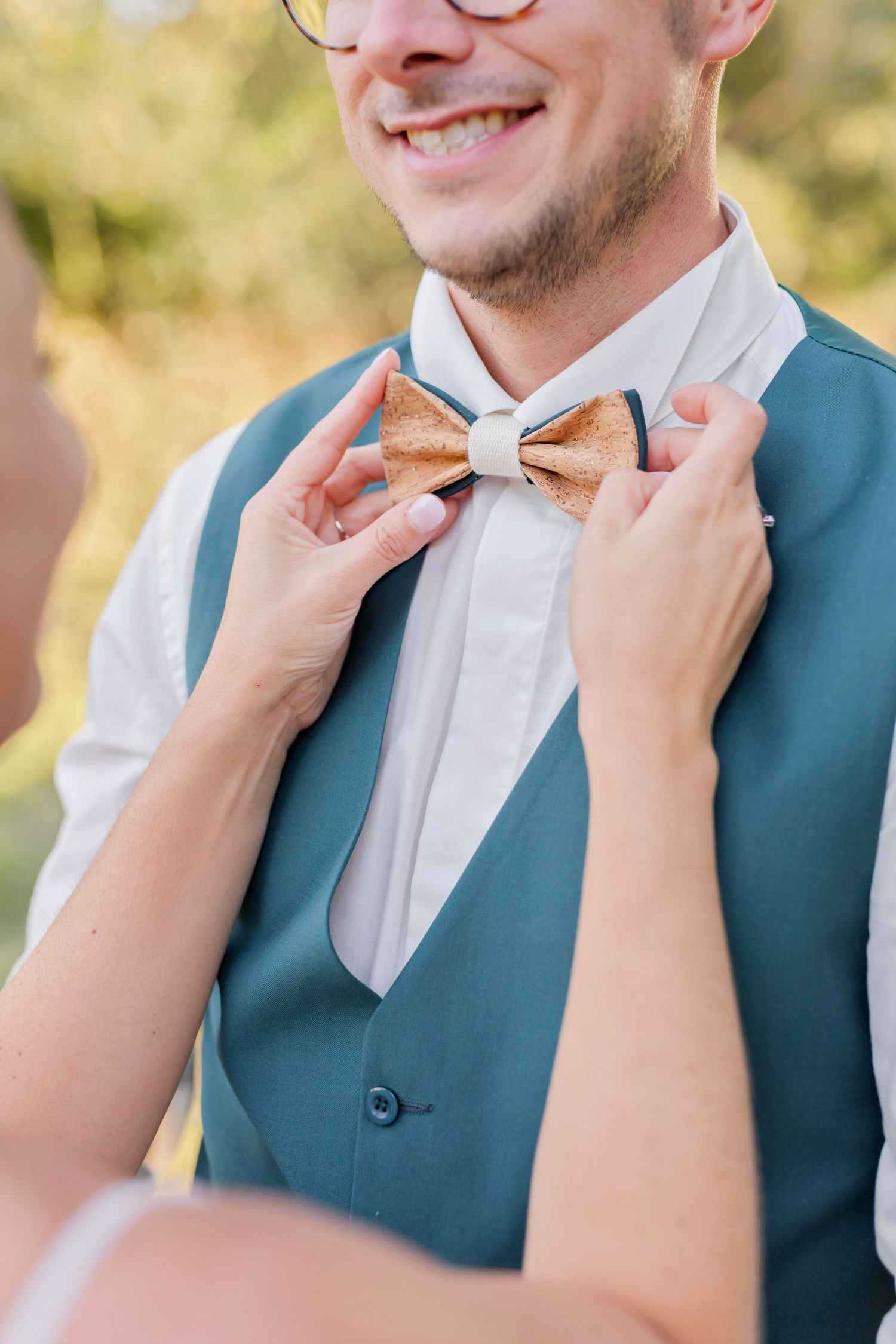 Hands adjust a tan bow tie on a teal vest and white dress shirt.