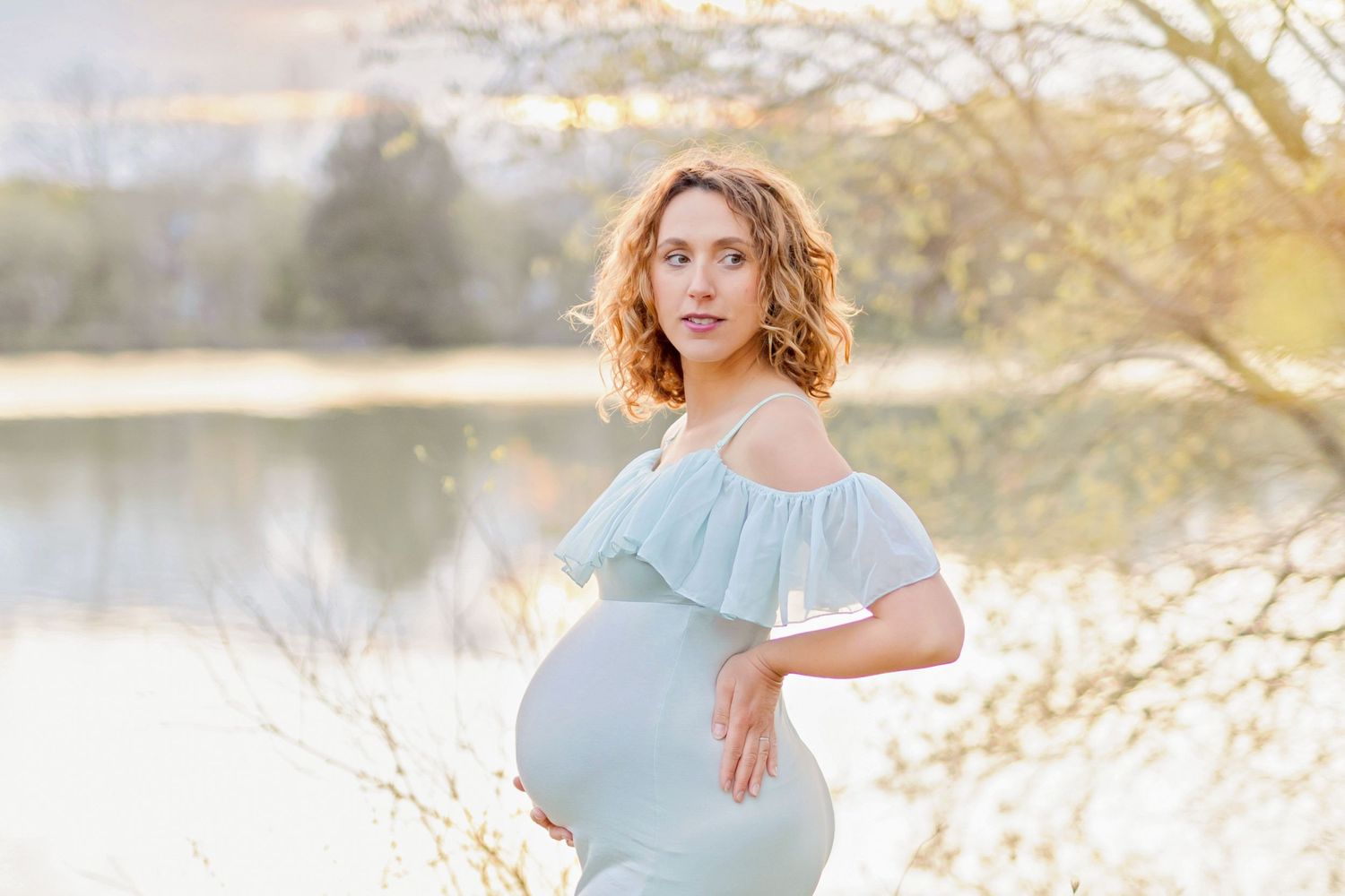 Dreamy lakeside maternity photo with a light blue mermaid-style dress at golden hour.