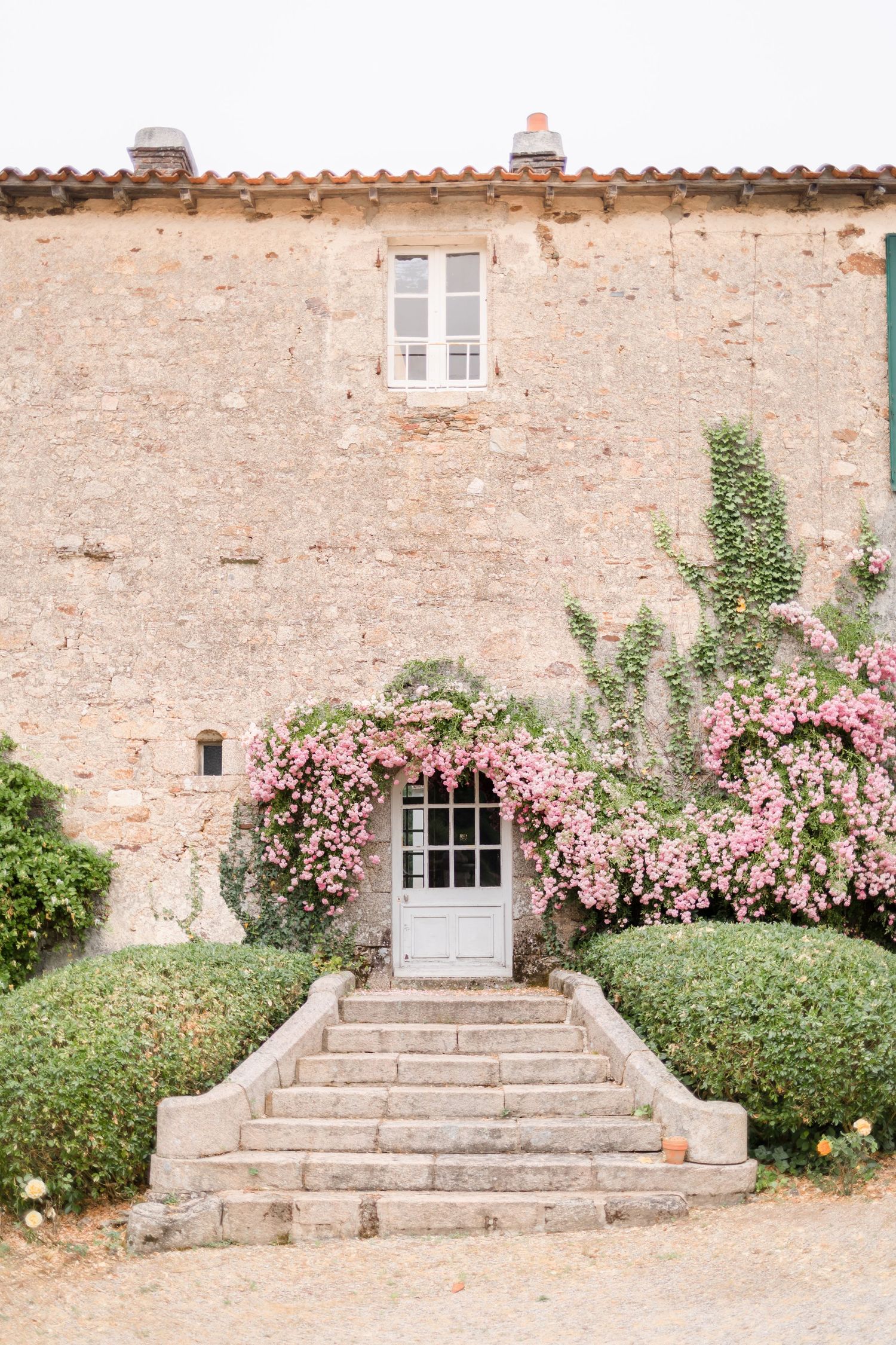 Stone facade of a French country house with pink climbing roses and stone steps leading to blue door.