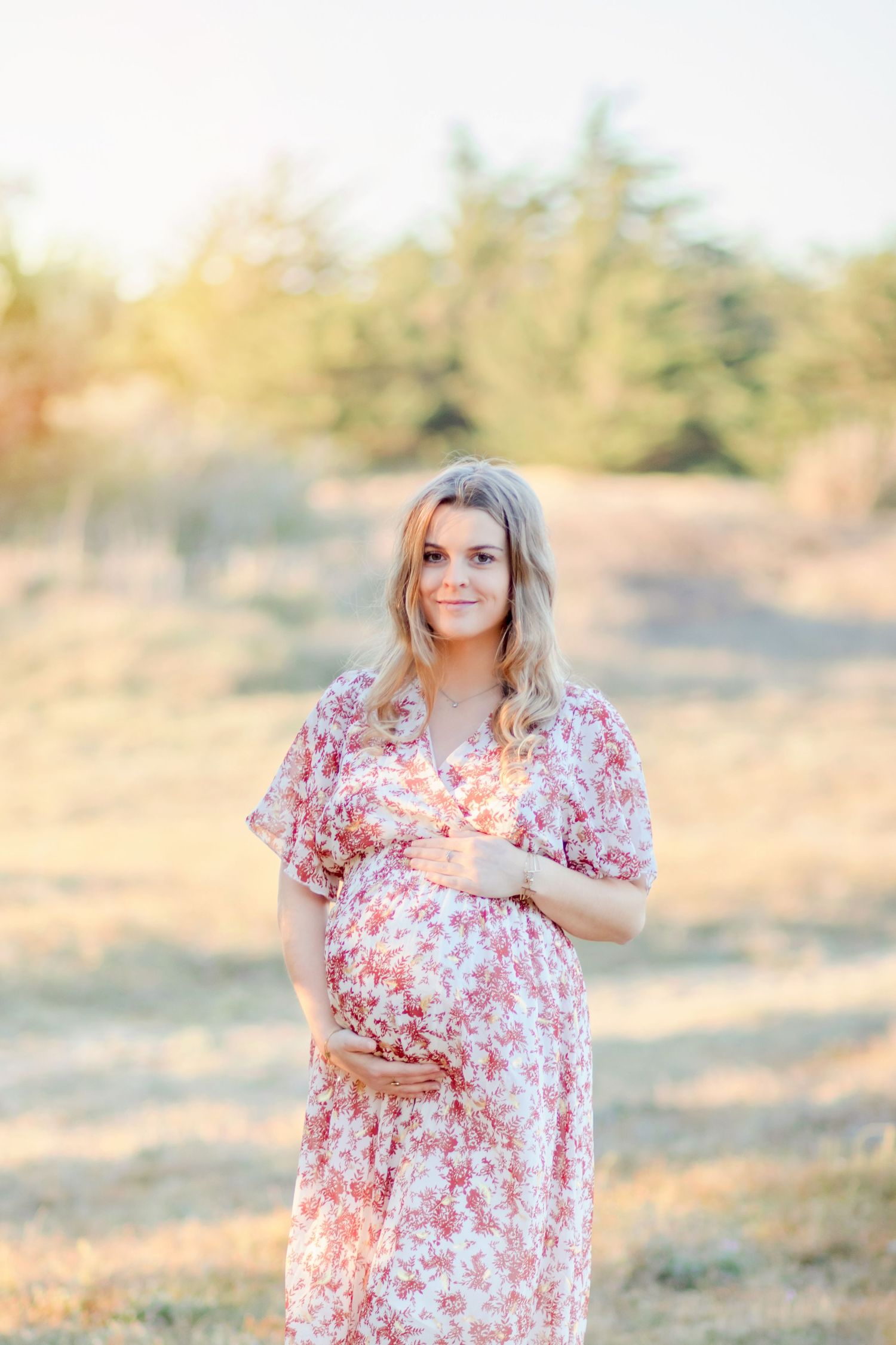 Expectant mother in floral pink dress poses in golden sunlit field.