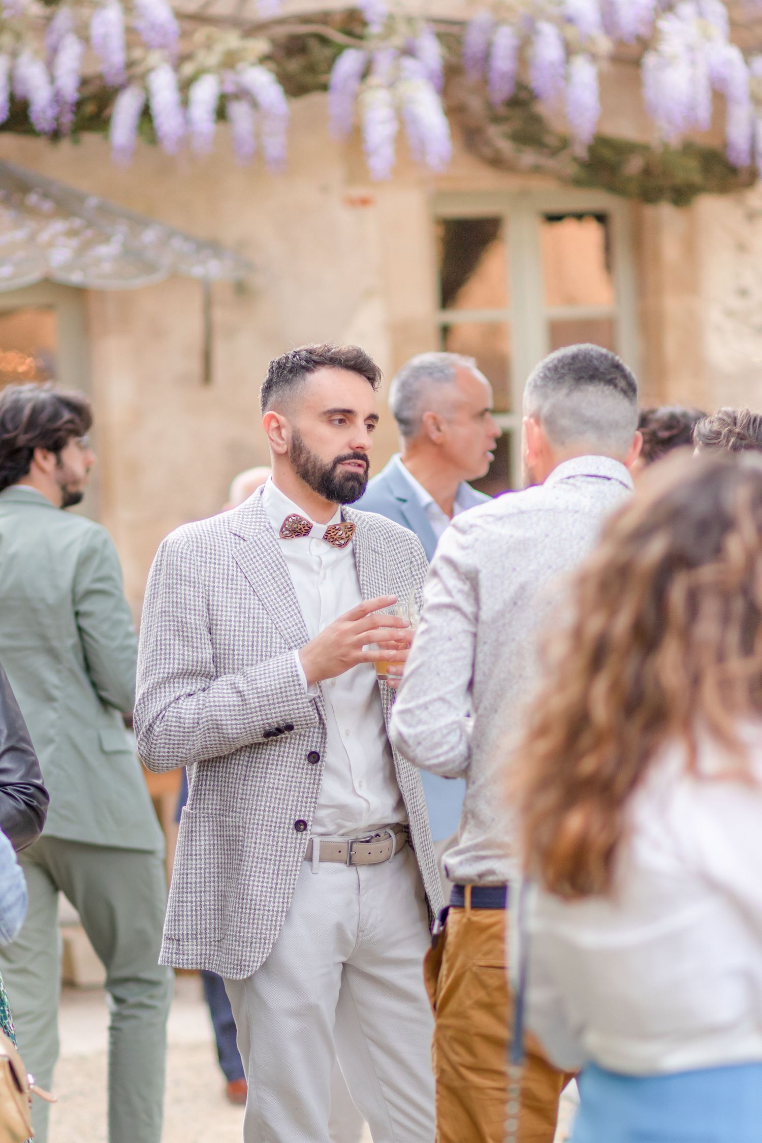 Elegant outdoor reception with guests mingling under purple wisteria blooms.