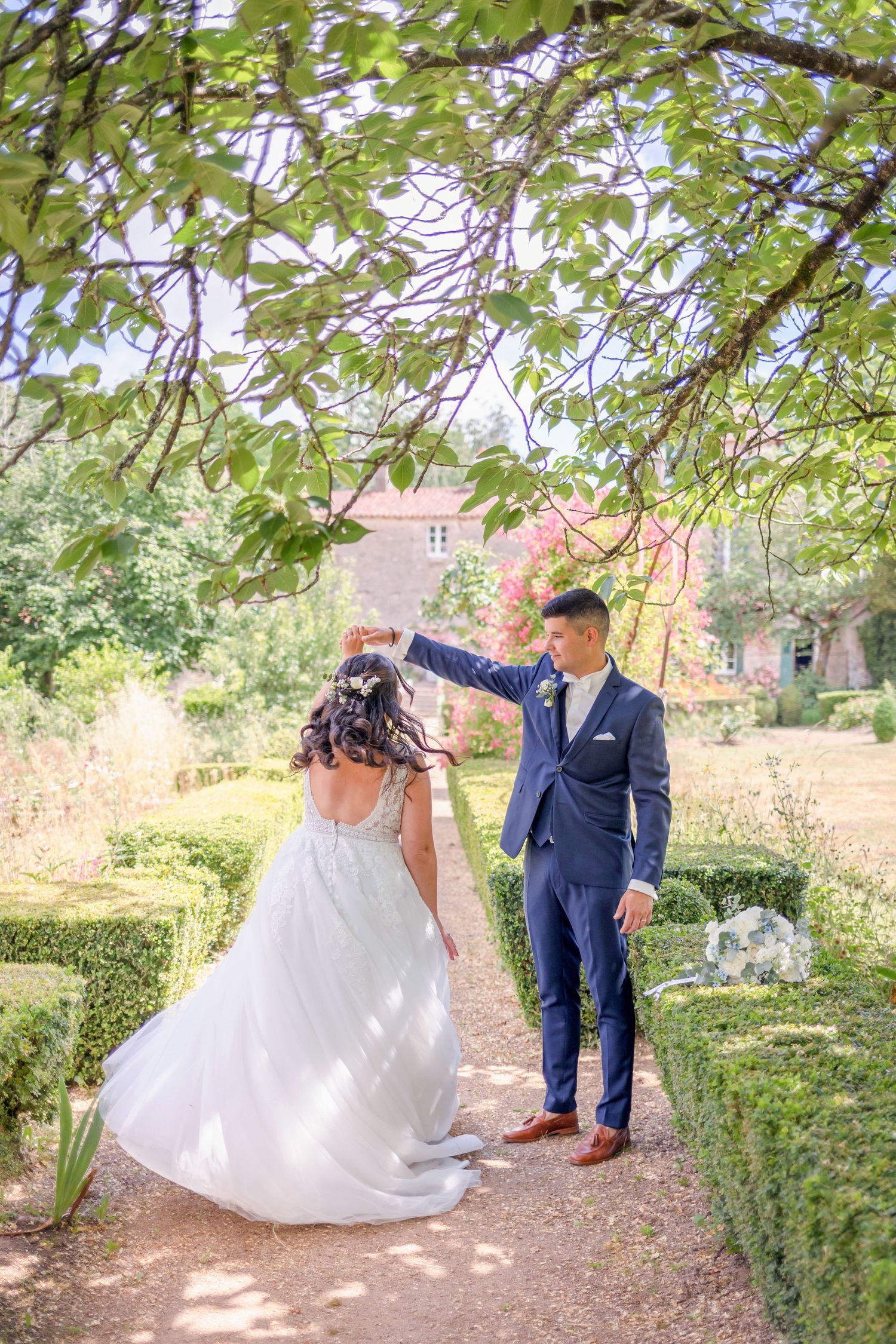 A garden wedding scene with a couple walking under leafy trees and hedges near a stone mansion.