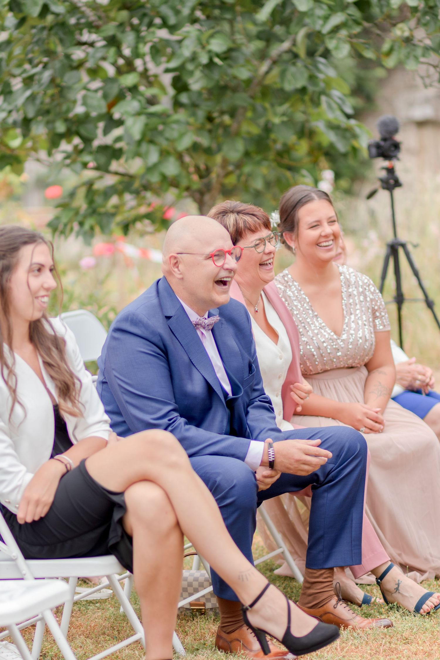 Wedding guests seated in white chairs outdoors laugh together during the ceremony.