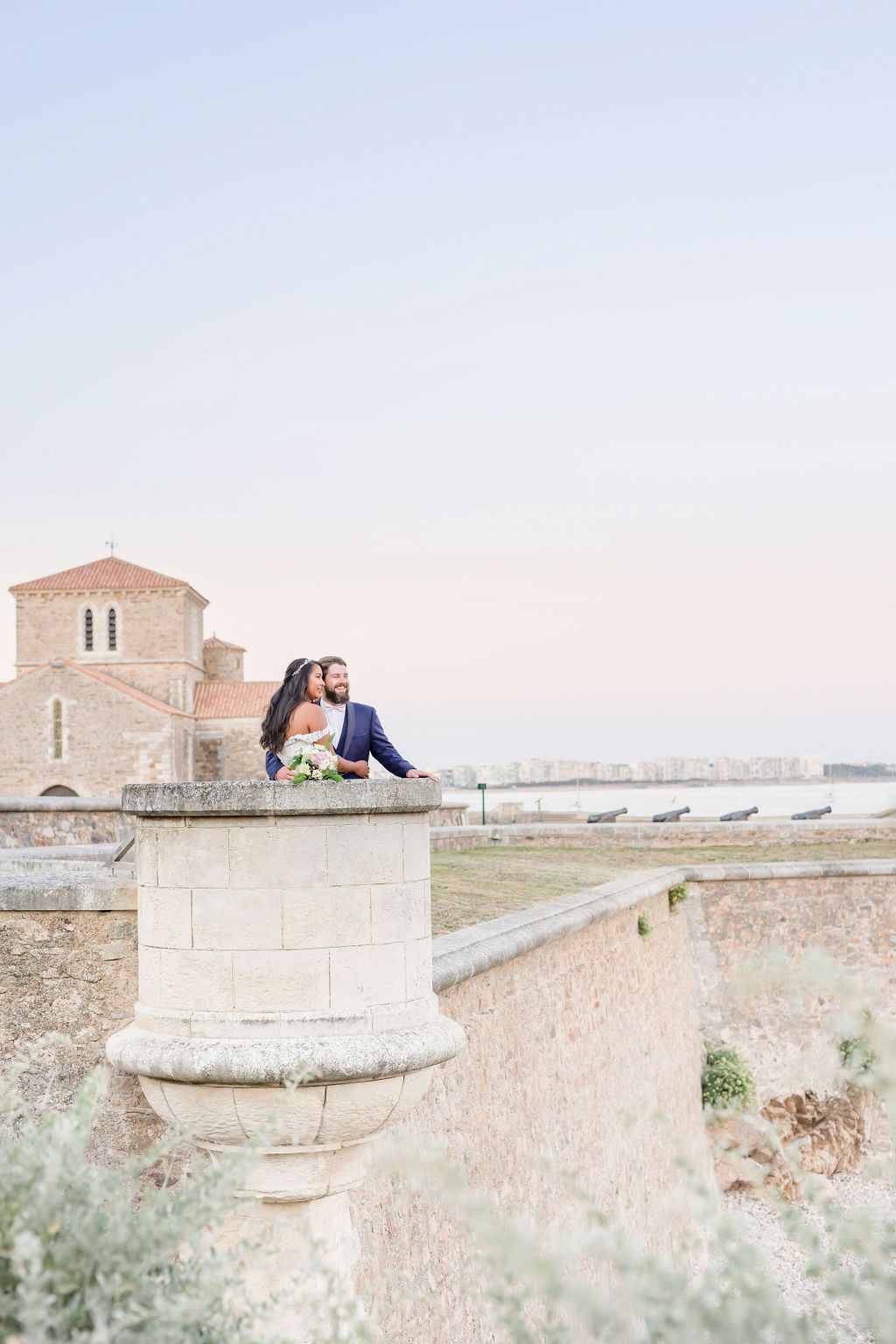 Couple sitting on historic stone pillar with coastal church and ocean views.