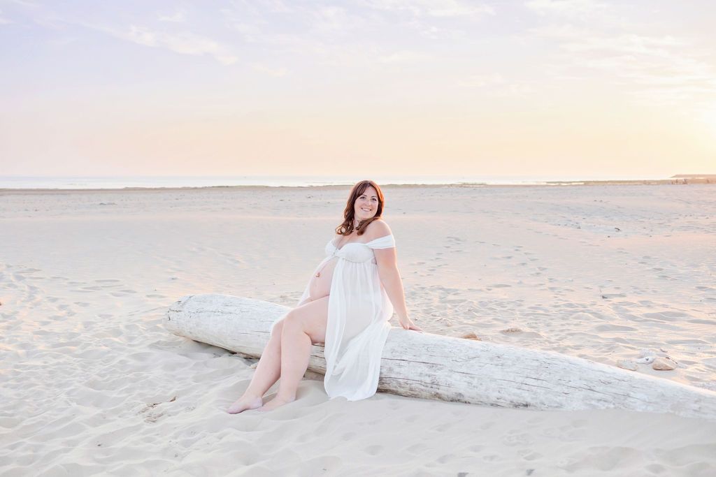 A scenic beach maternity photo session with flowing white dress and driftwood at sunset.