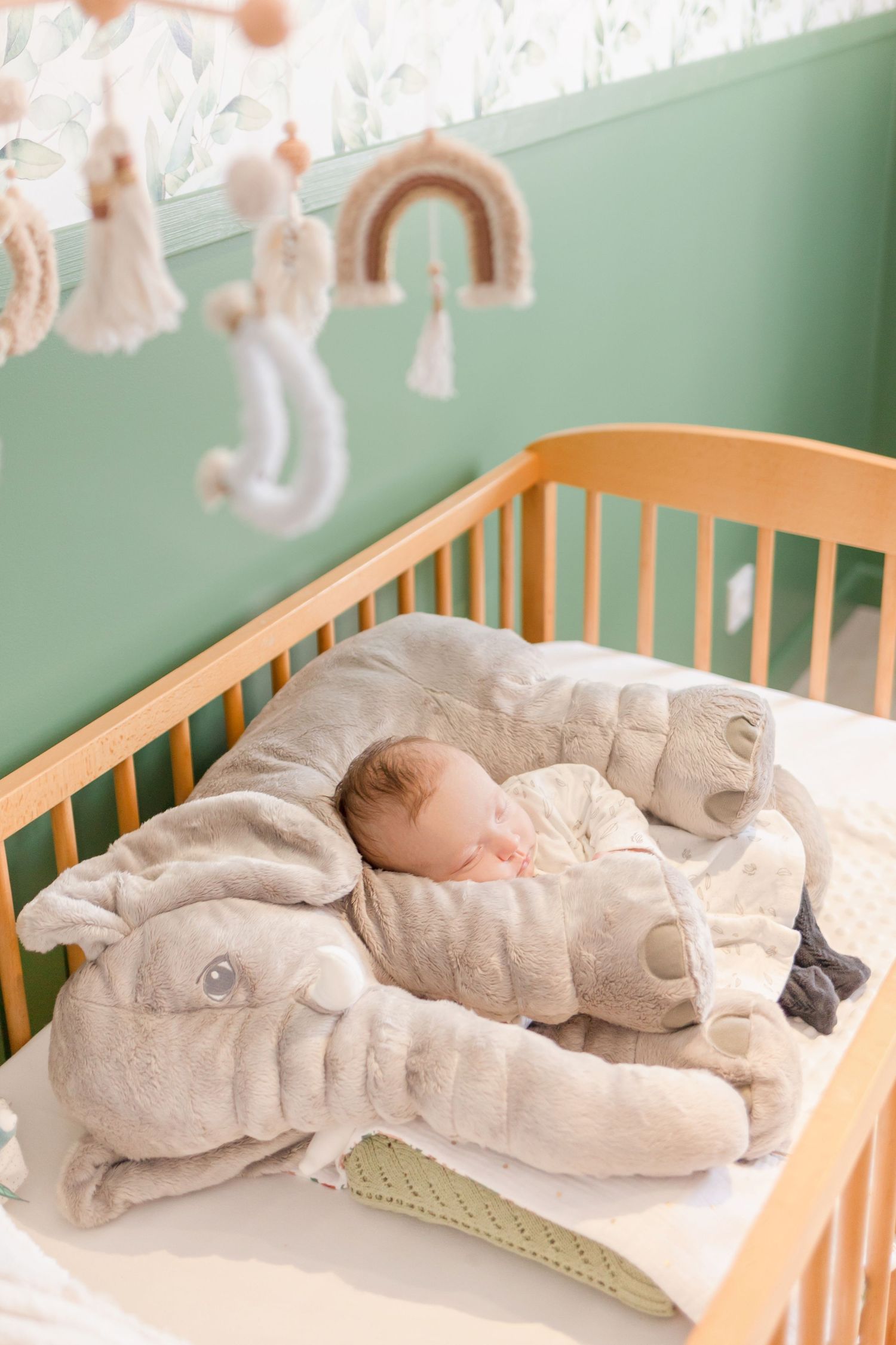 Sleeping infant nestled between gray elephant cushions in wooden crib under boho nursery decorations.
