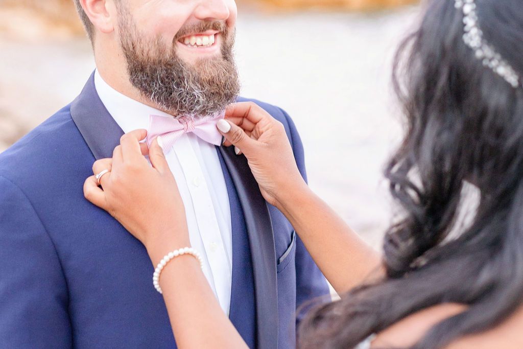 Hands adjust a pink bowtie on a navy blue suit jacket during a wedding photoshoot.