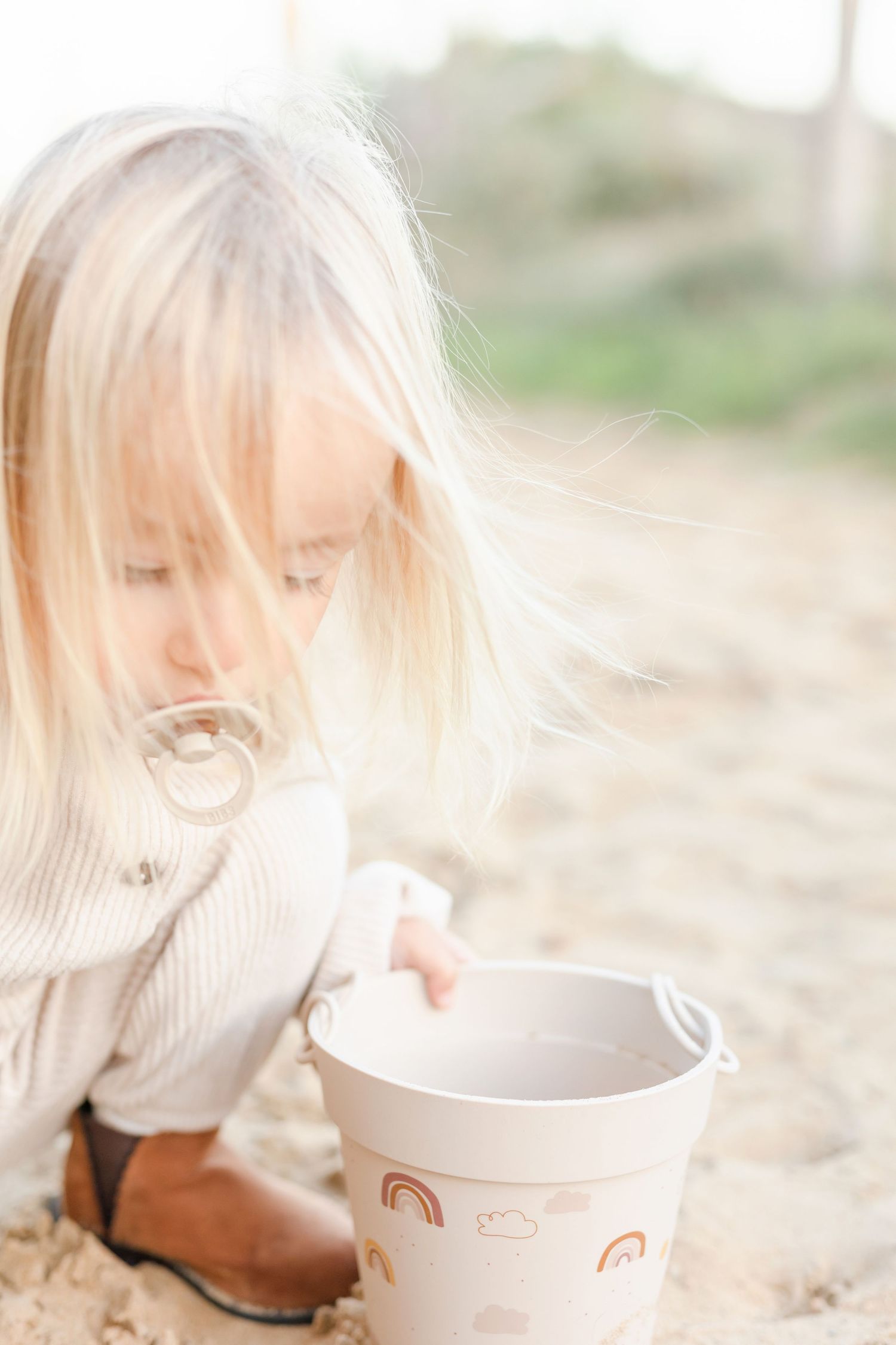 Young child in cream sweater plays with decorative bucket on sandy ground.
