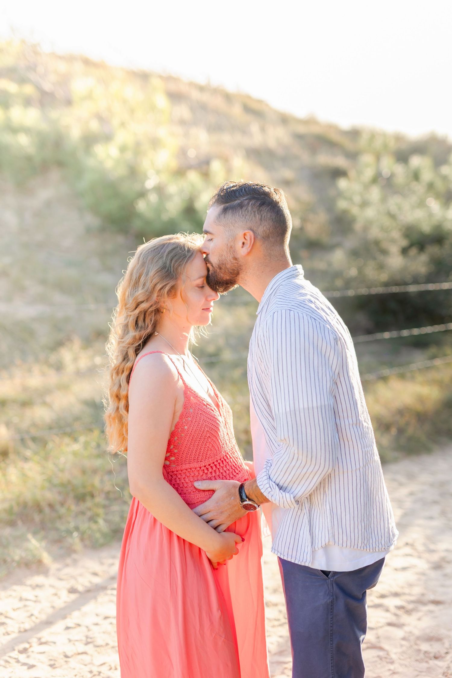 A couple shares an intimate moment during a sunset maternity photoshoot, with her wearing a coral dress.
