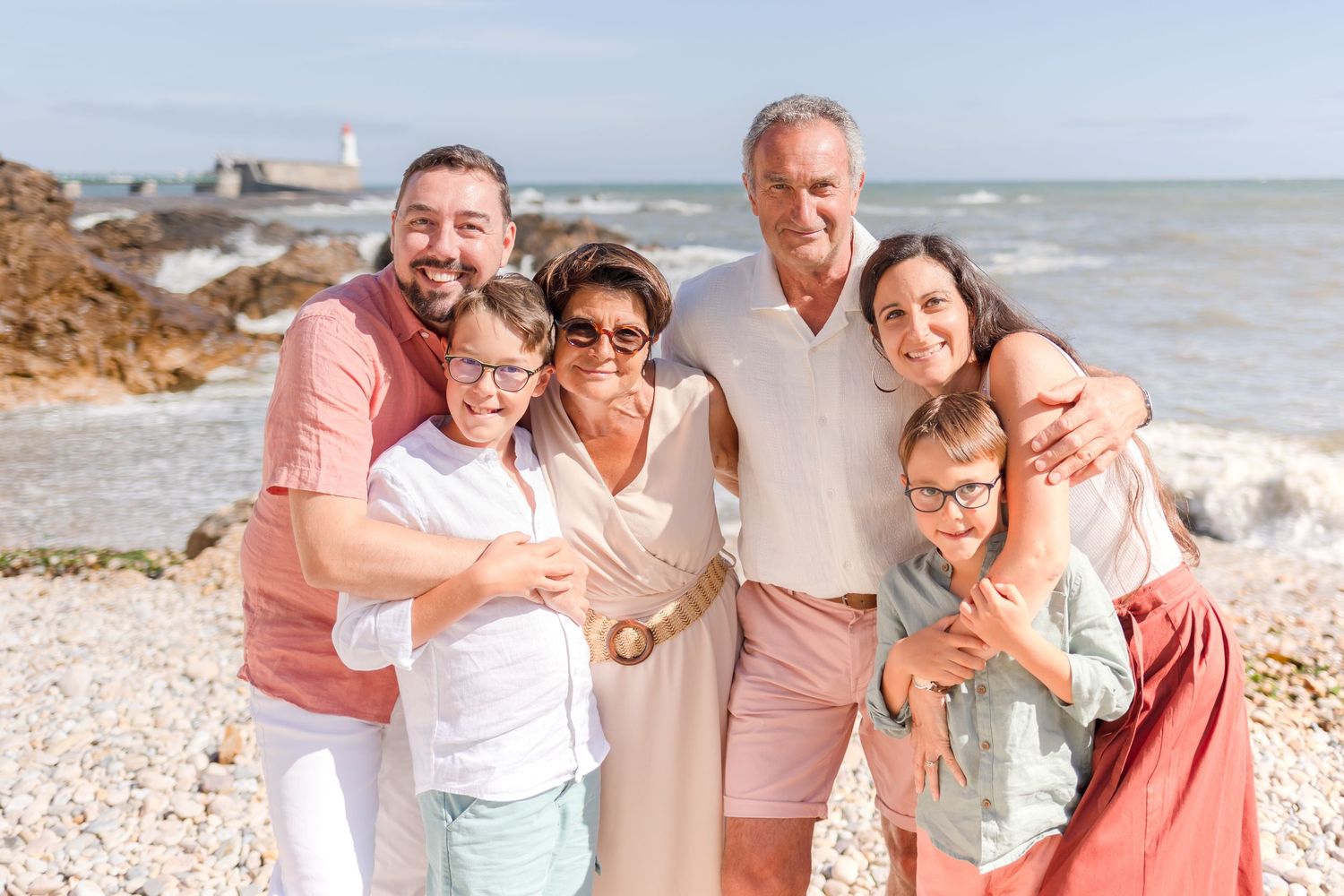 A family embraces while posing for a photo on a rocky beach with ocean waves in the background.
