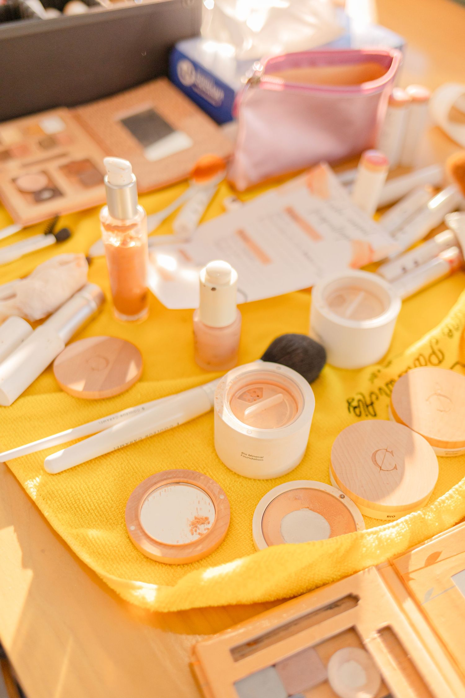 Makeup products and cosmetics arranged on yellow cloth surface.