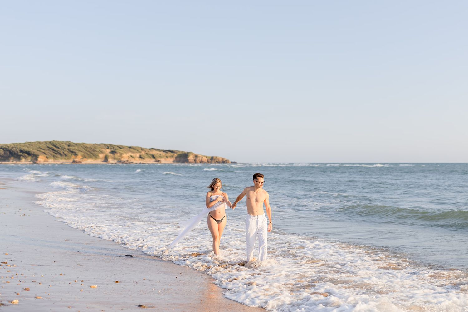 A couple walks together along a sandy beach shoreline with cliffs in the background on a sunny day.