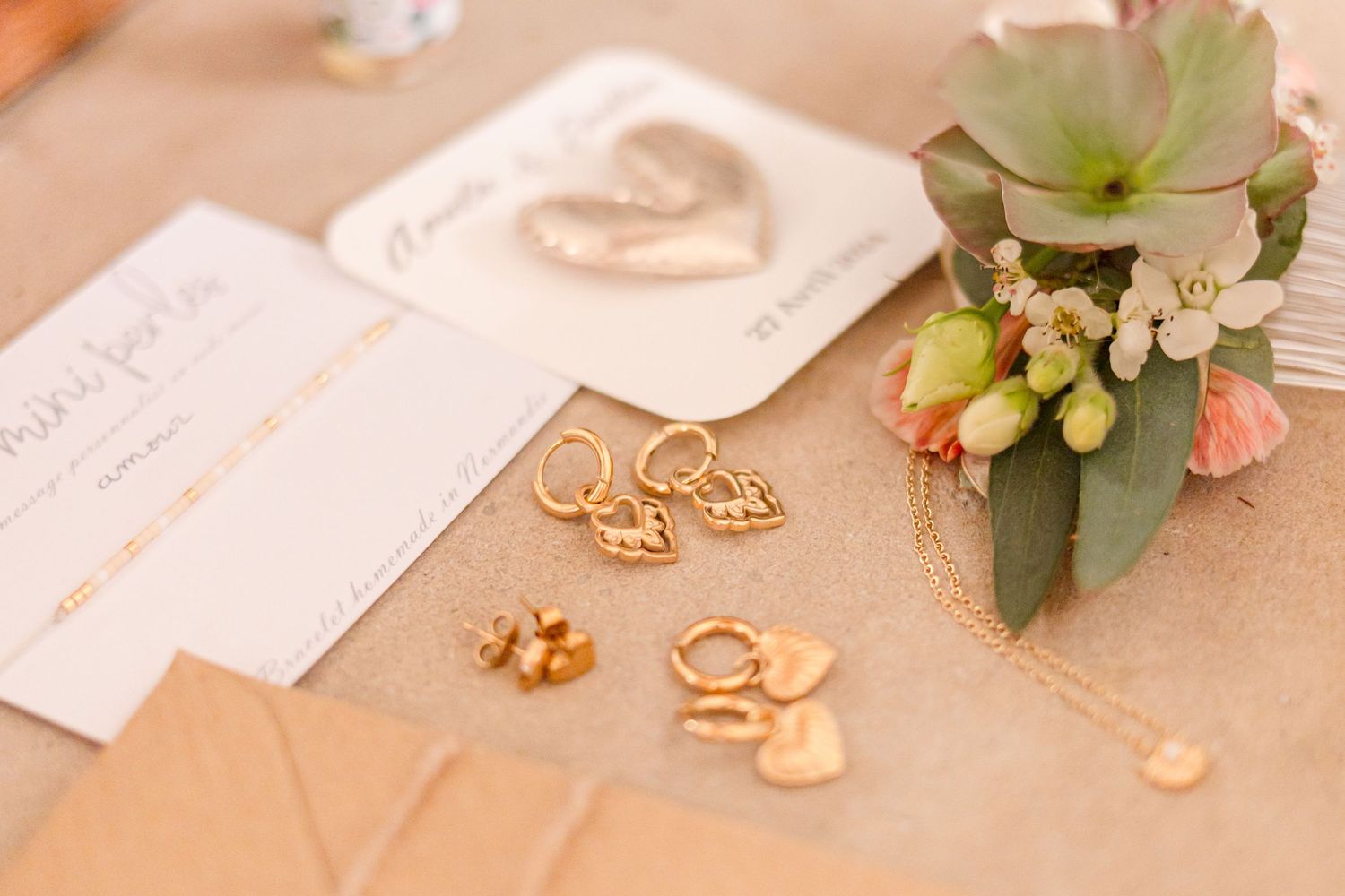 Wedding details including gold jewelry, cards with hearts, and a small succulent arrangement on a beige surface.