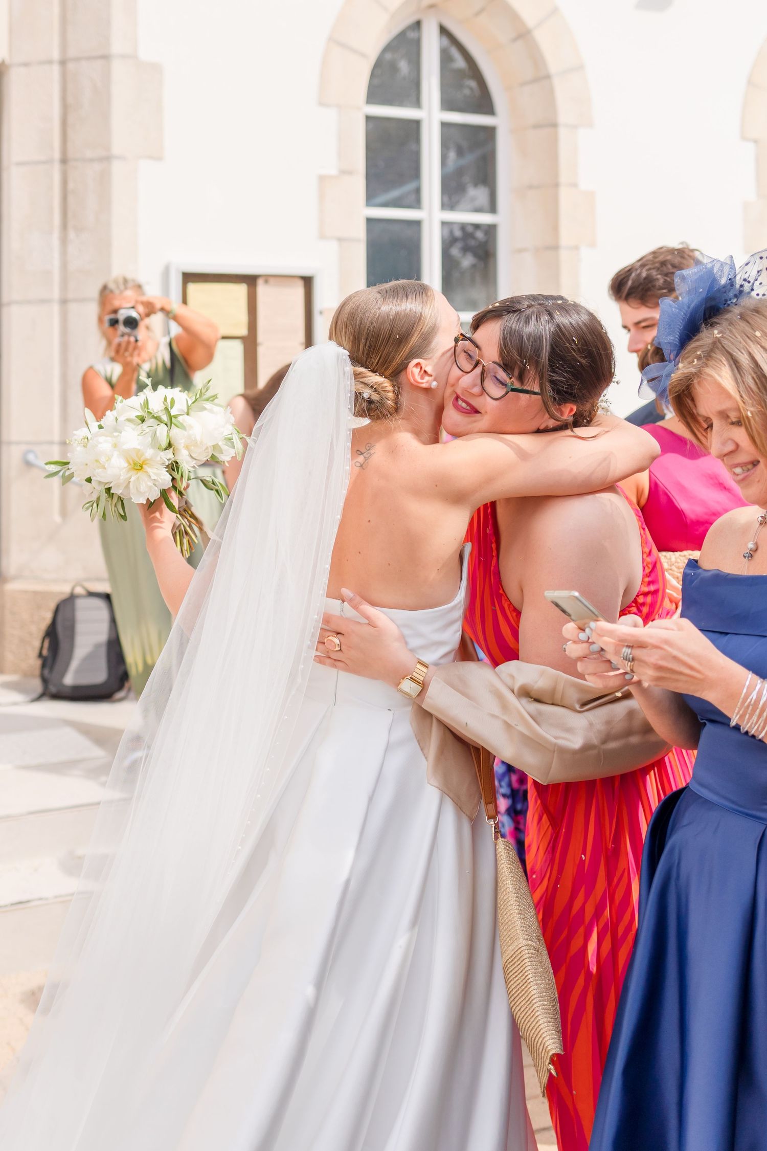 Wedding guests share warm embraces and congratulations outside a church with arched windows.