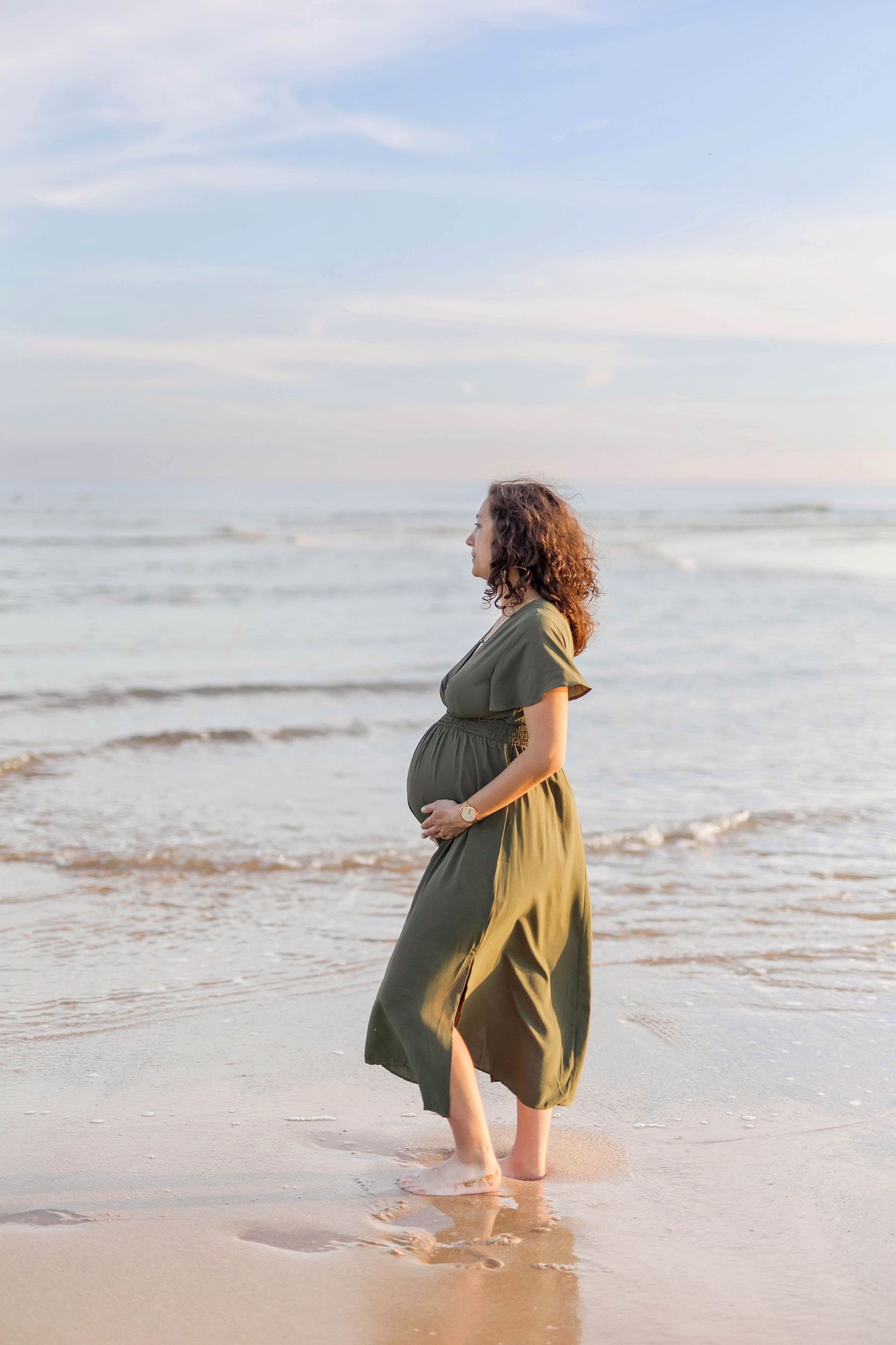 A person in a green dress stands at the ocean's edge during sunset, silhouetted against the waves.