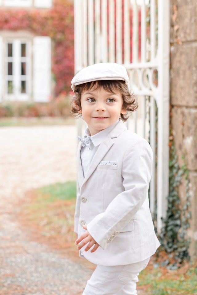 Child in white formal attire and cap stands in front of stone columns.