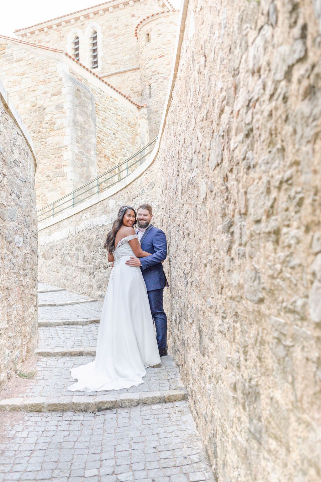 Wedding couple embracing on cobblestone path beside historic stone wall.
