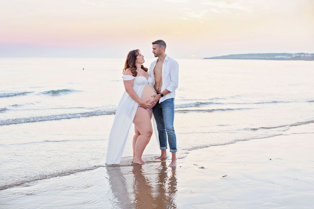 A romantic beach maternity photography session during golden hour with waves crashing in the background.