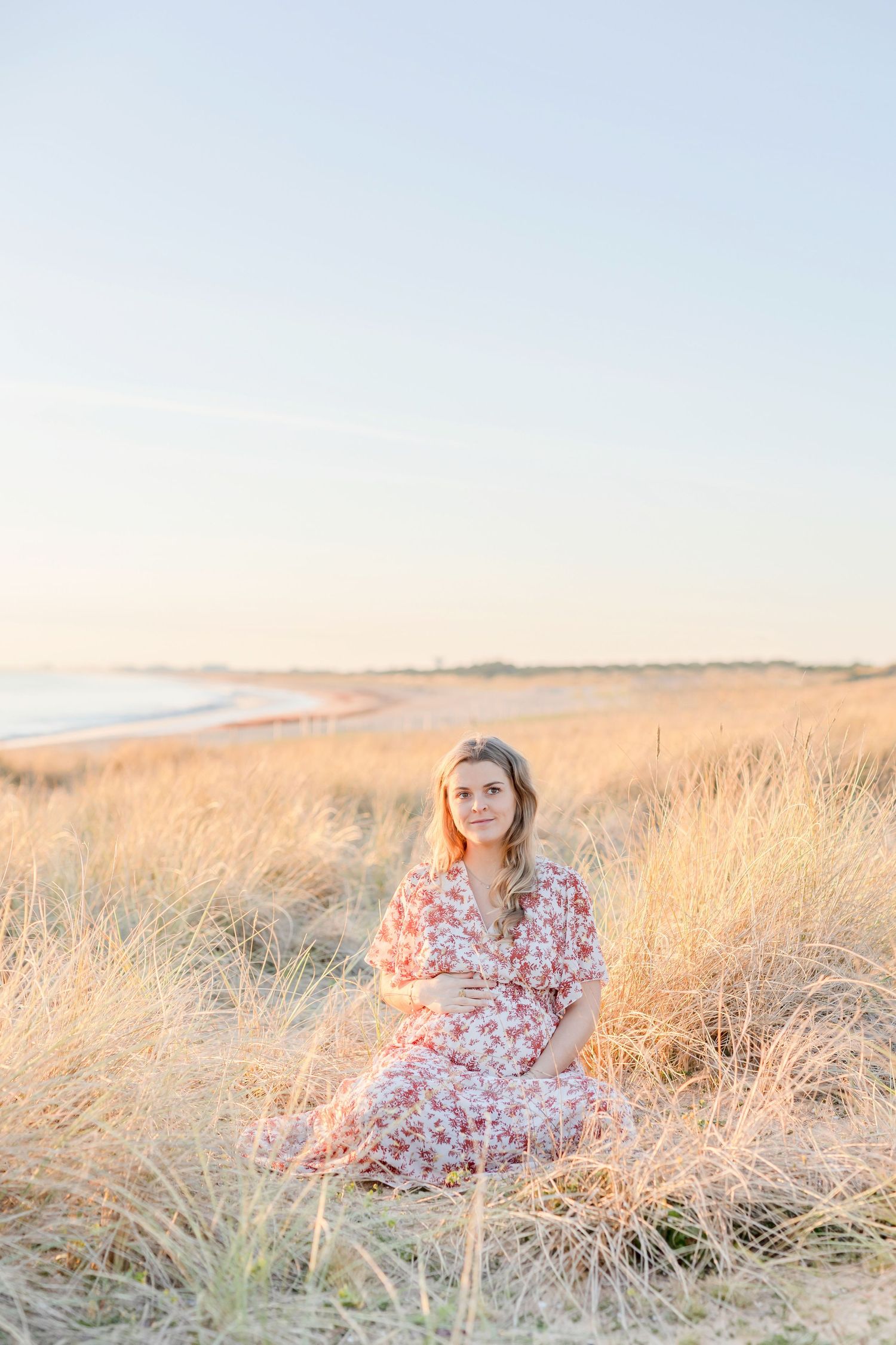 Woman in floral dress sitting among tall coastal grasses at sunset.
