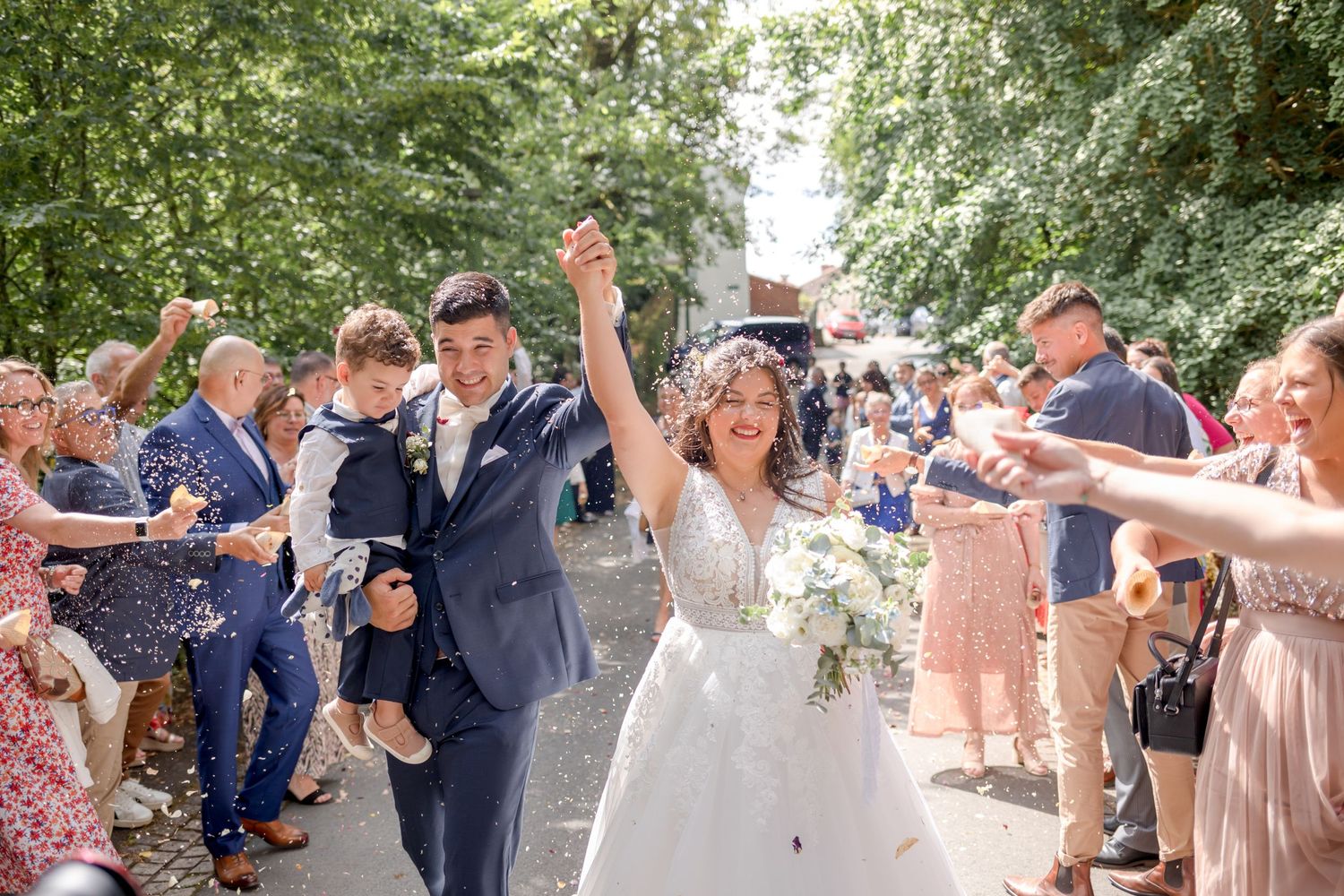 Wedding guests throw confetti at newlyweds walking down a tree-lined path in celebration.