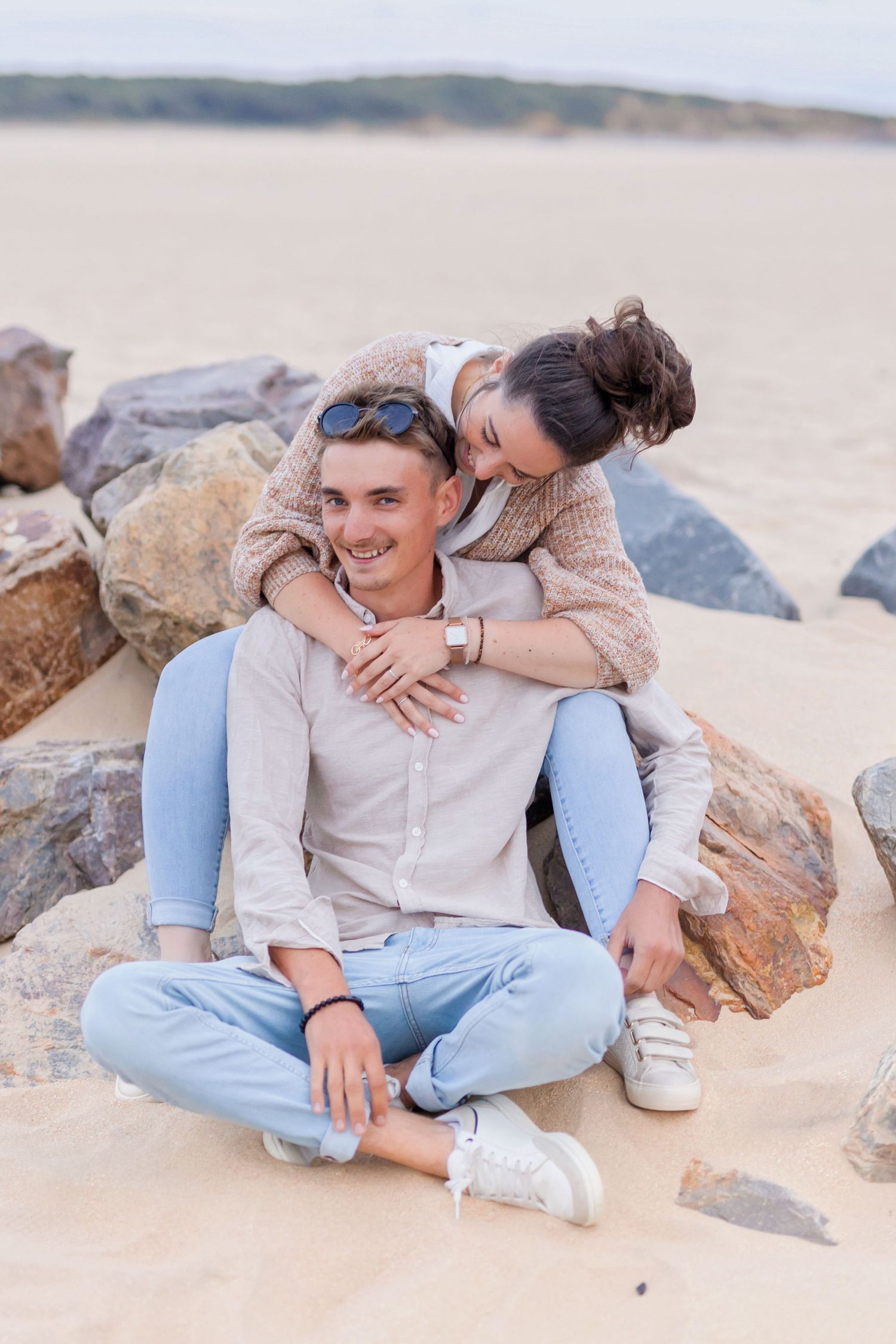 Playful couple embraces while sitting on sandy beach near rocks on a cloudy day.
