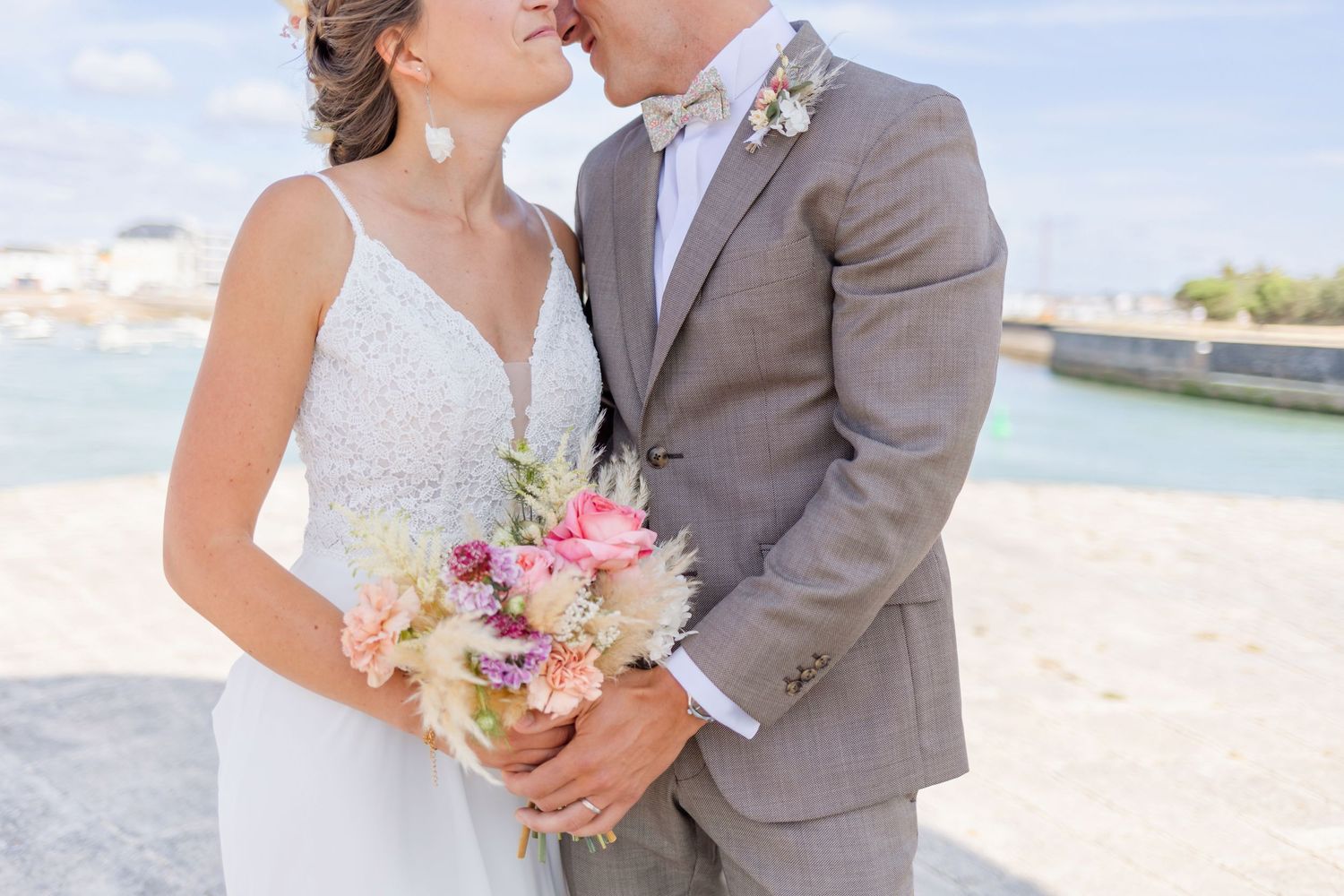 Close up of wedding couple with pink floral bouquet near coastal lighthouse.