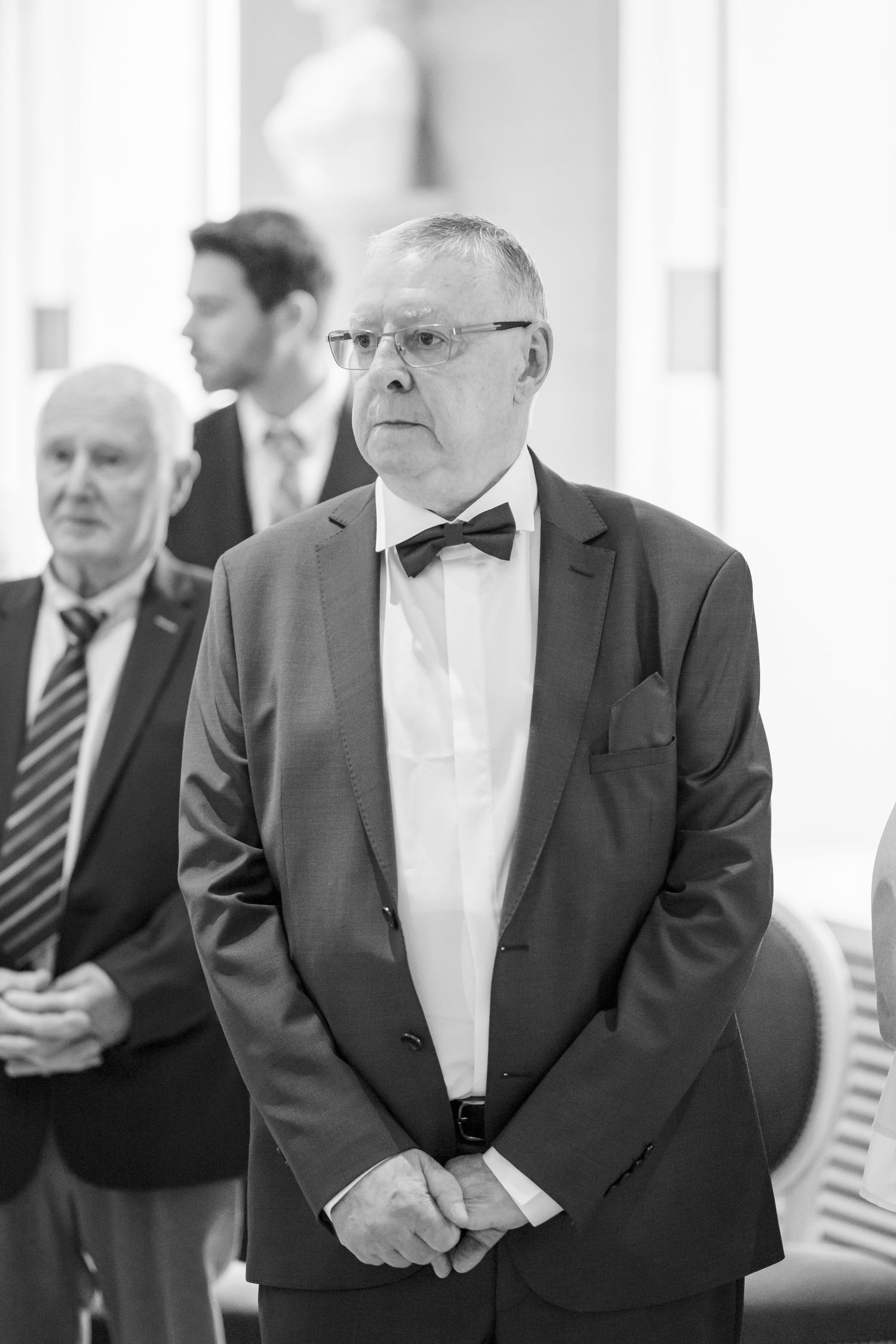 Formal wedding guests in black tuxedos and bow ties gather during elegant reception.