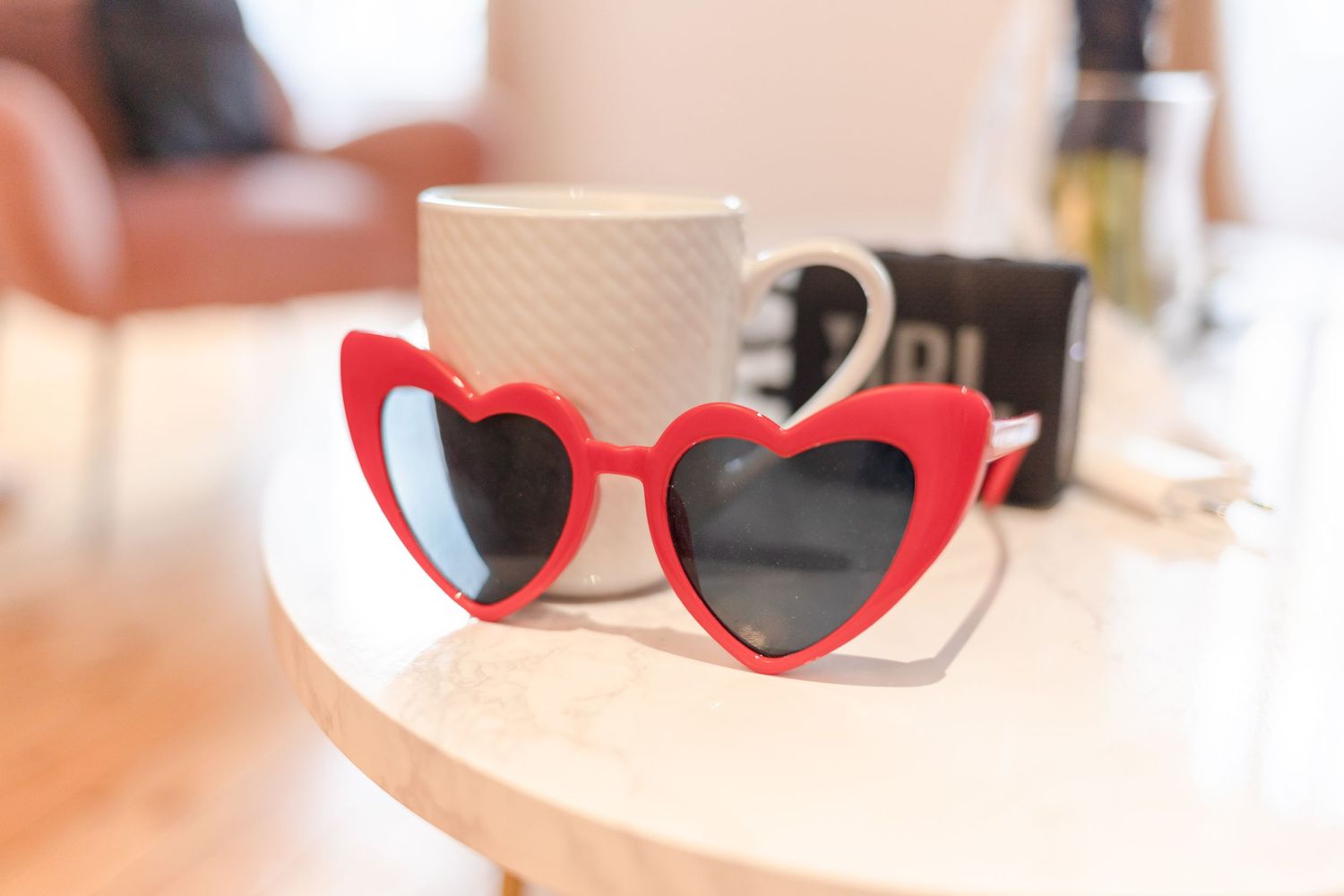 Red heart-shaped sunglasses next to white coffee mug on table.