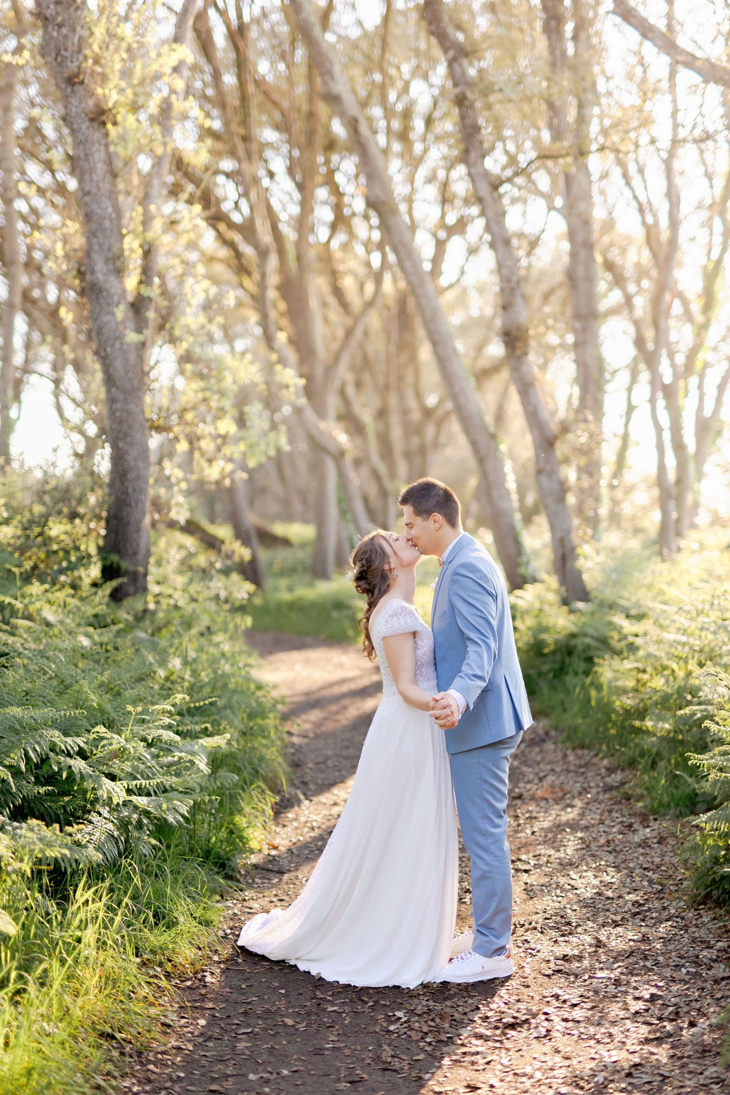 A couple in wedding attire shares a tender moment on a forest path surrounded by ferns.