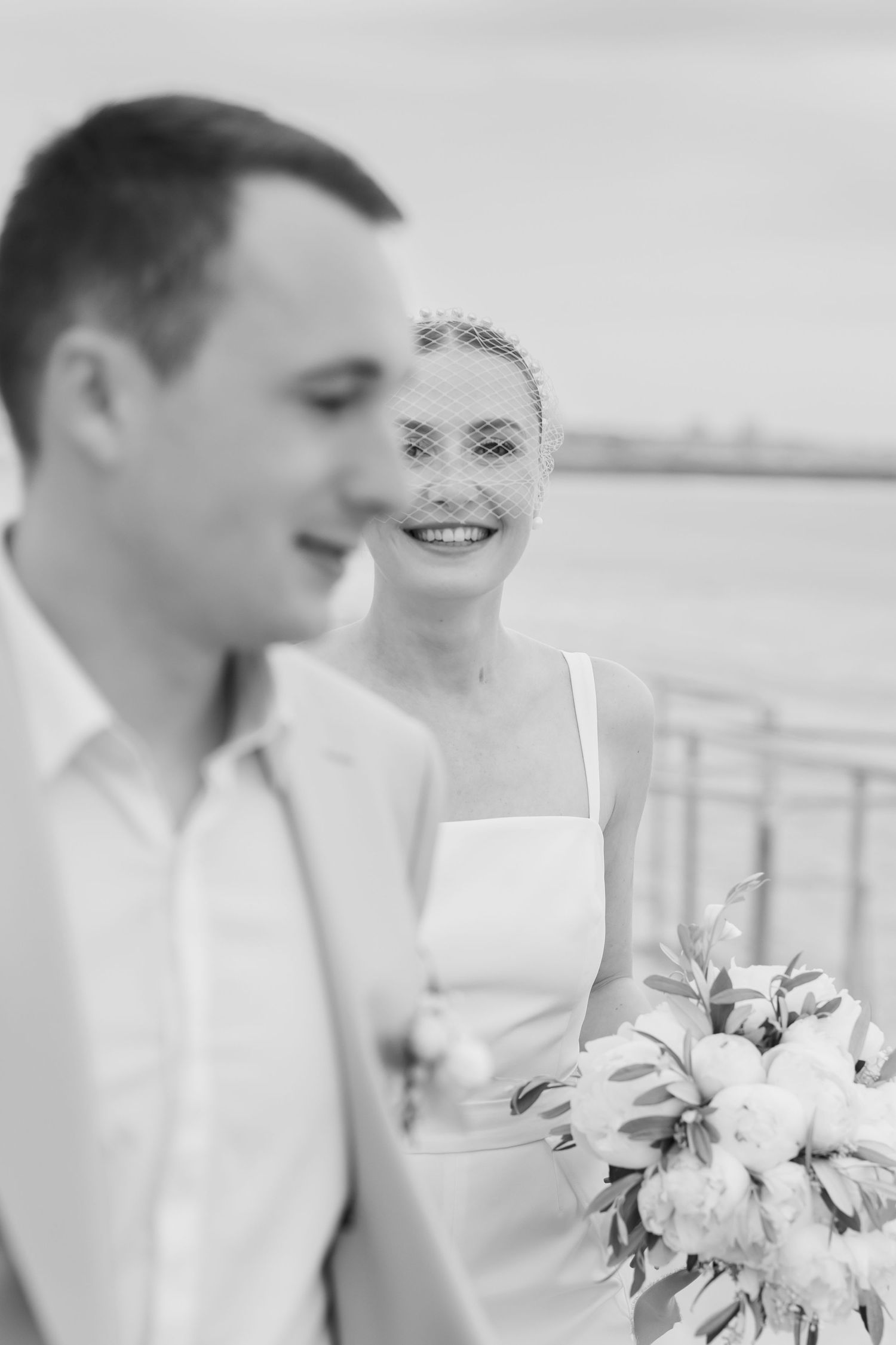 A romantic black and white wedding portrait showing a couple by the water with a bridal bouquet.