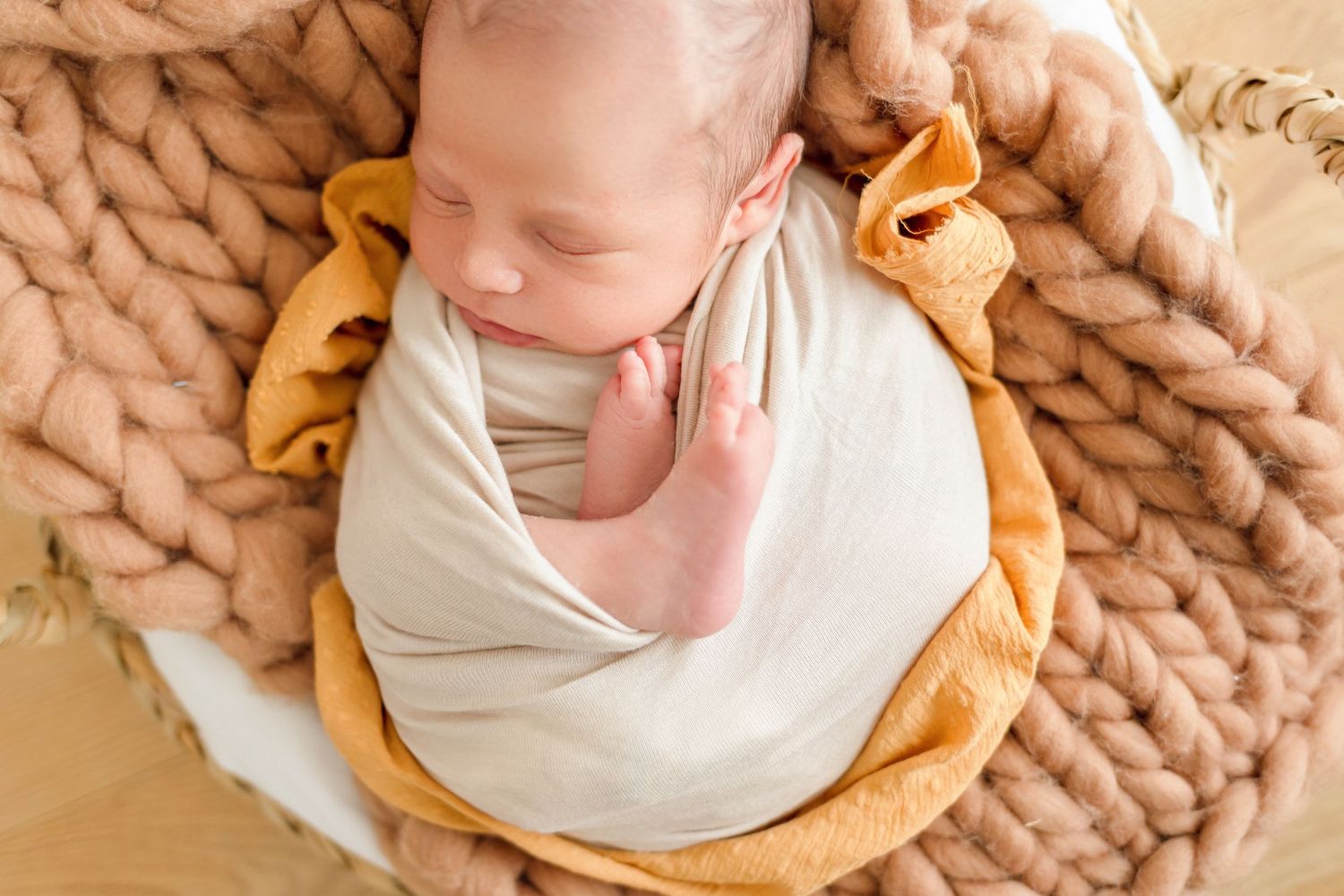 Newborn baby wrapped in cream fabric sleeping on chunky knit blanket with orange accents.
