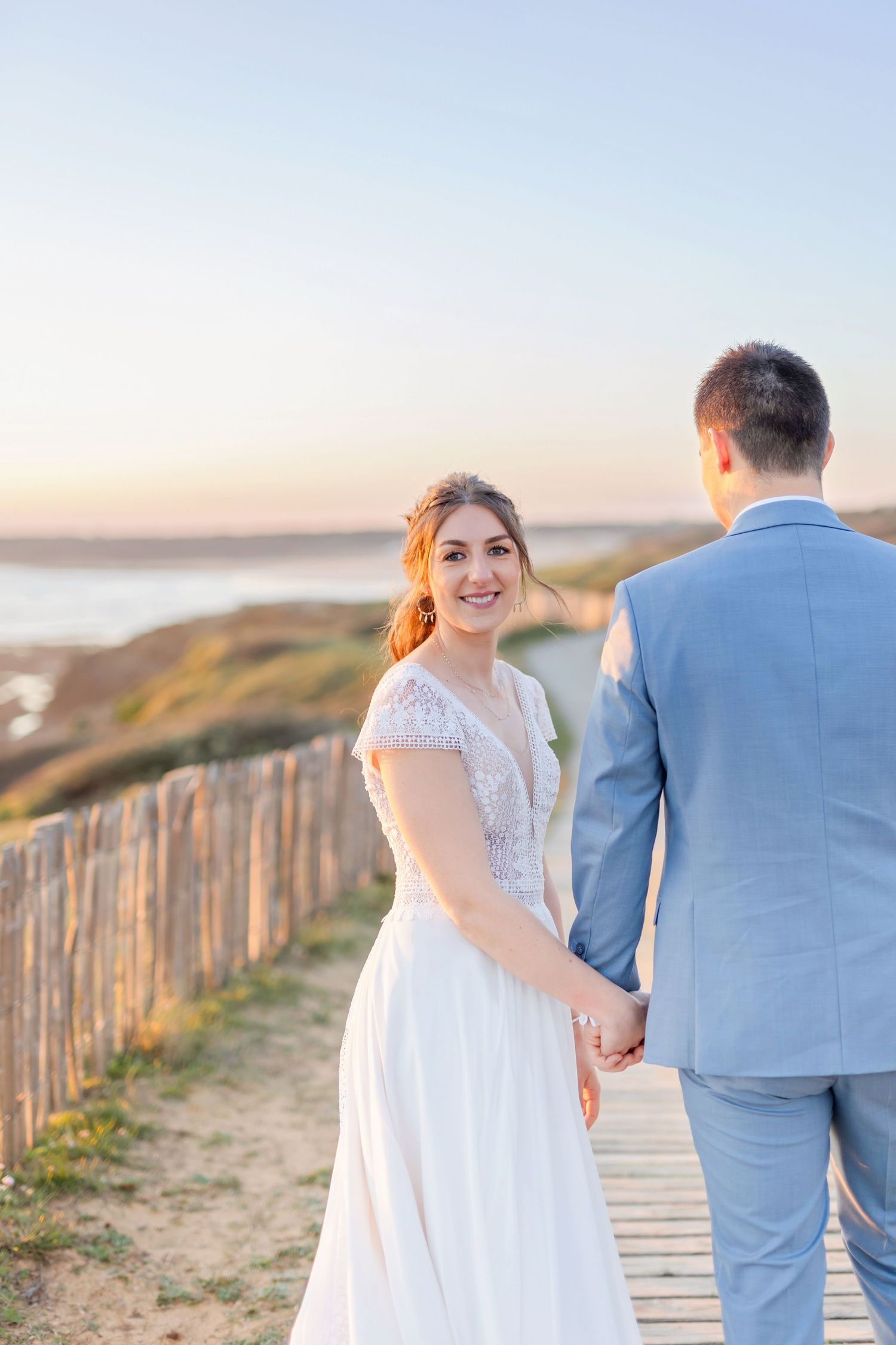 Wedding couple holding hands on coastal boardwalk during golden sunset with ocean views.